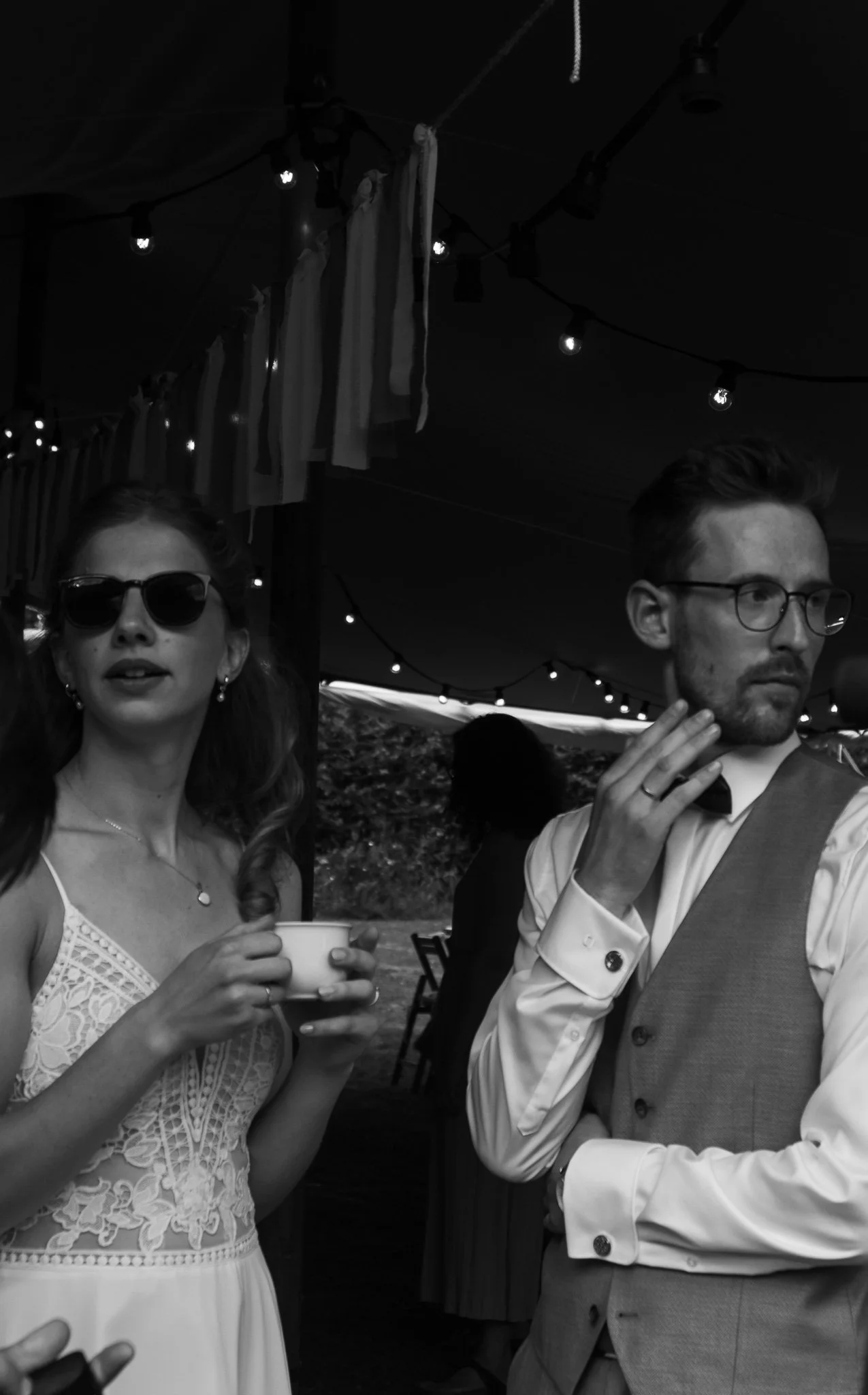 Zwei Menschen bei einer Party im Freien, eine Frau trägt Sonnenbrille und hält eine Tasse, ein Mann trägt eine Weste und Brille, beide unter einer festlichen Beleuchtung.