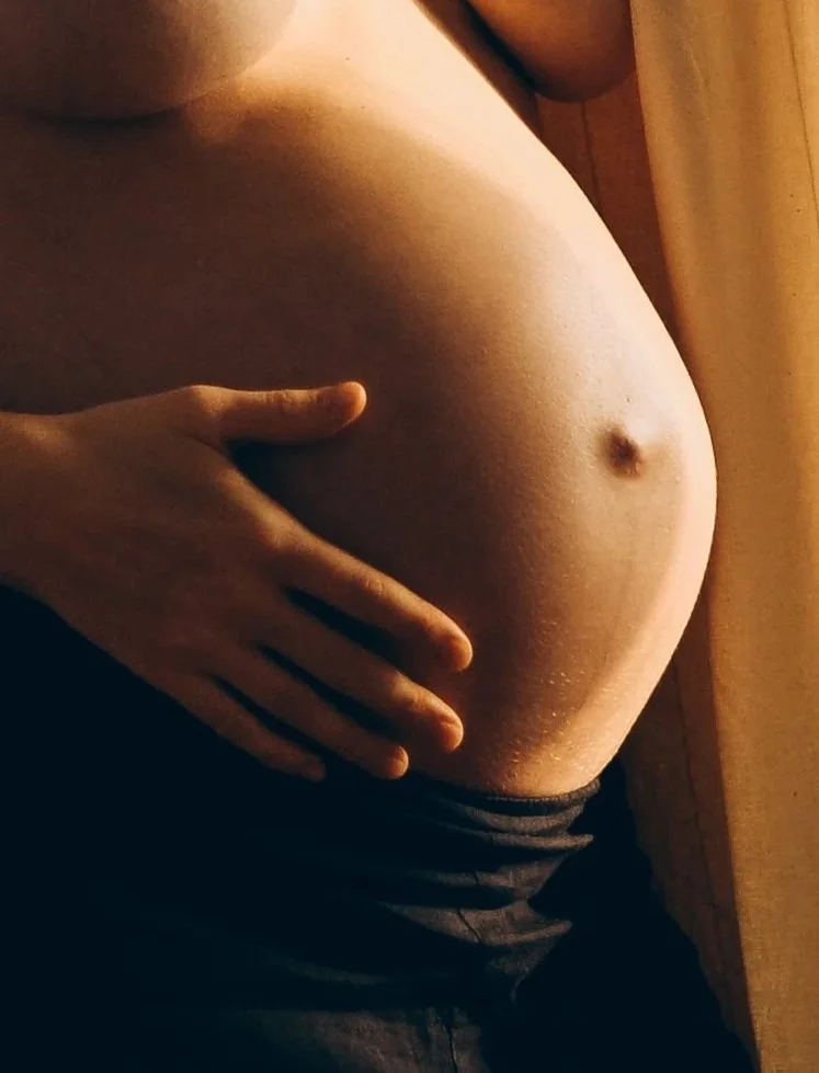 Nackte Frau, die ihren Bauch mit der Hand hält, in einer natürlichen Umgebung, mit warmem Licht und einer dunklen Hose.