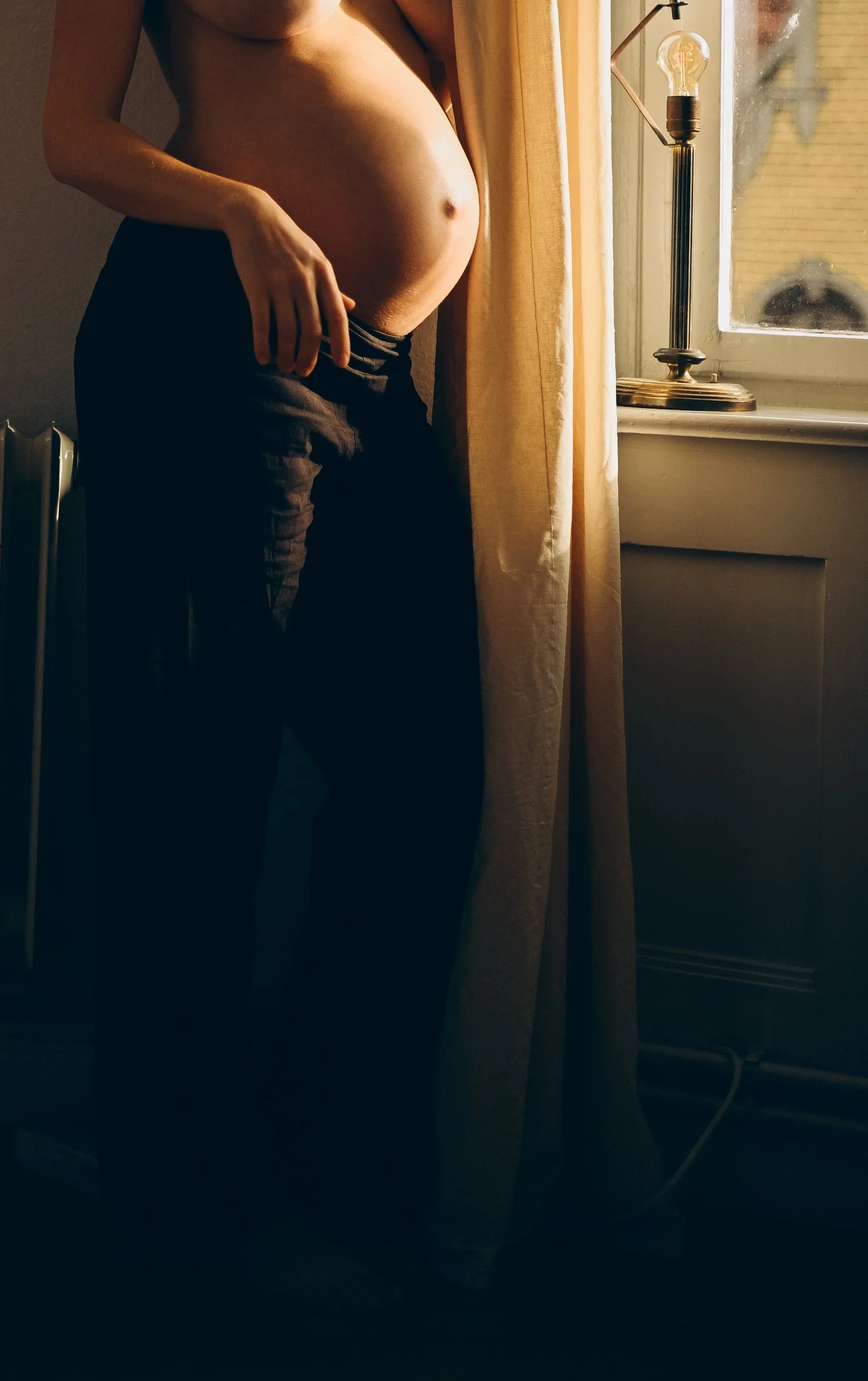Eine schwangere Frau steht bei Sonnenuntergang in einem Raum neben einem Fenster mit gelben Vorhängen, sie ist nackt von der Taille aufwärts.