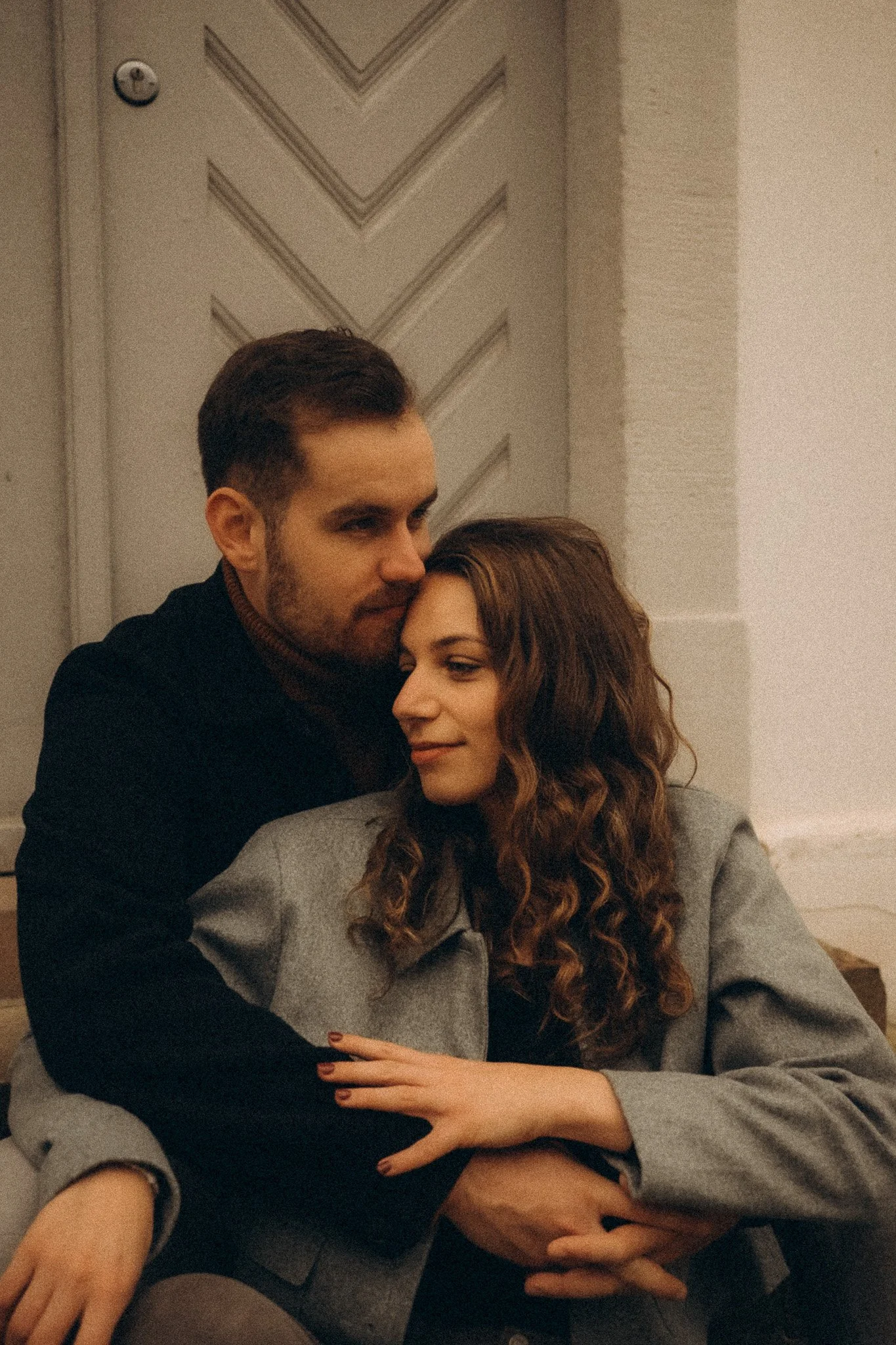Ein Mann und eine Frau sitzen eng zusammen auf einem Sofa. Der Mann umarmt die Frau und die beiden wirken glücklich und verliebt. Der Hintergrund ist eine helle Wand mit einem dezenten Muster.
