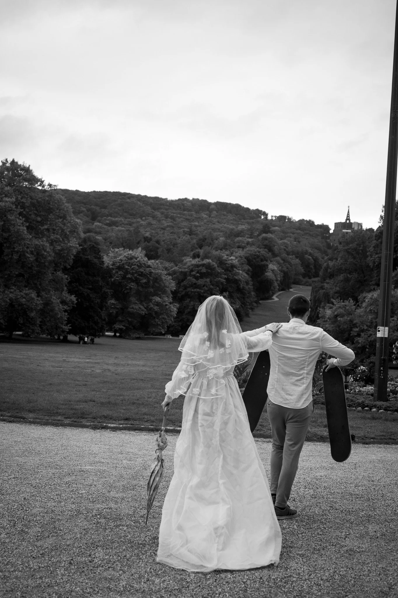 Paare in Hochzeitskleidung spazieren im Park, die Frau trägt ein Brautkleid und hält einen Regenschirm, der Mann trägt ein Hemd und hält Skates.