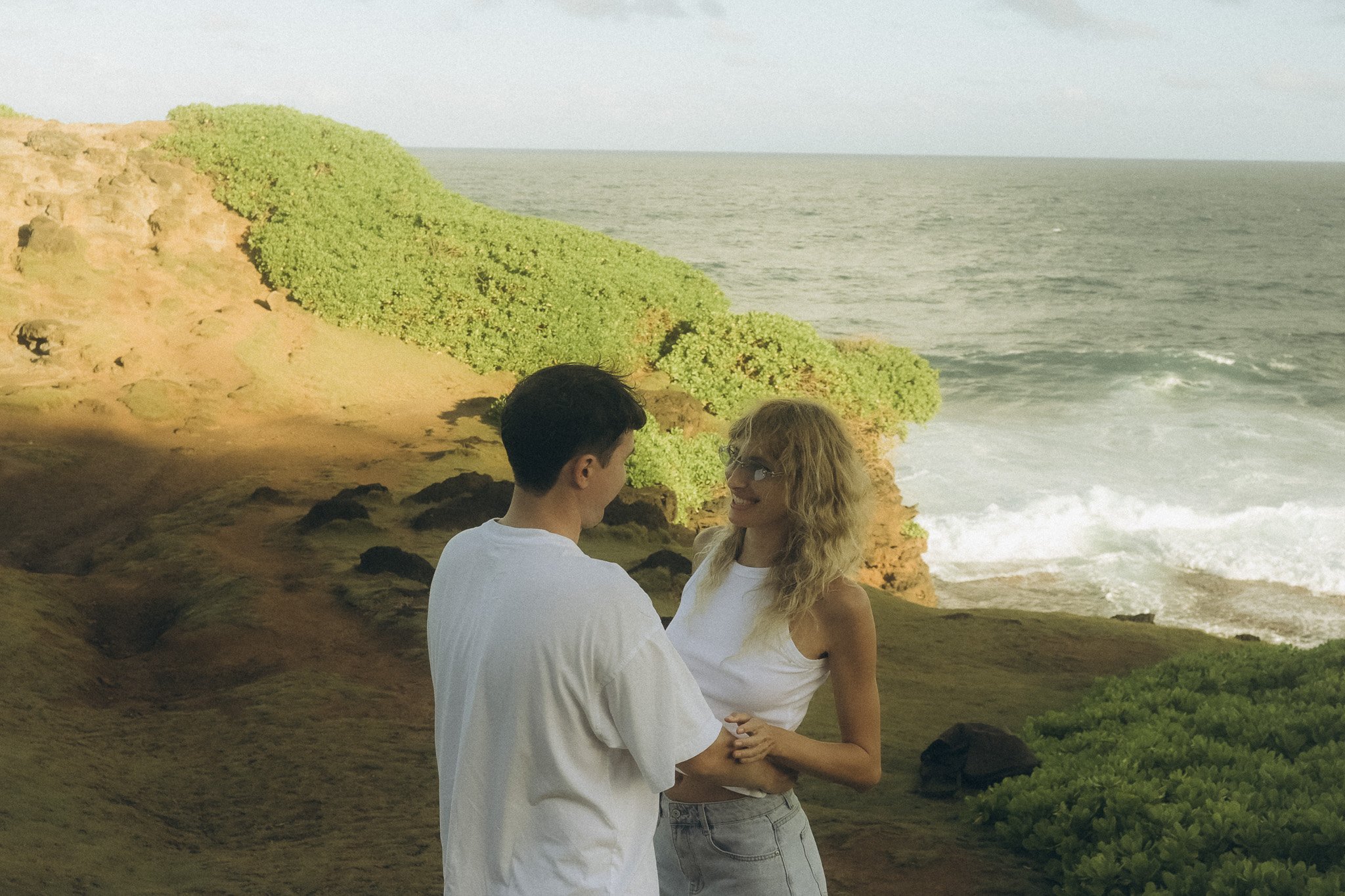 Ein Mann und eine Frau stehen sich an einer Klippe am Meer gegenüber, halten sich an den Händen und schauen sich an, während die Wellen gegen die Felsen schlagen.