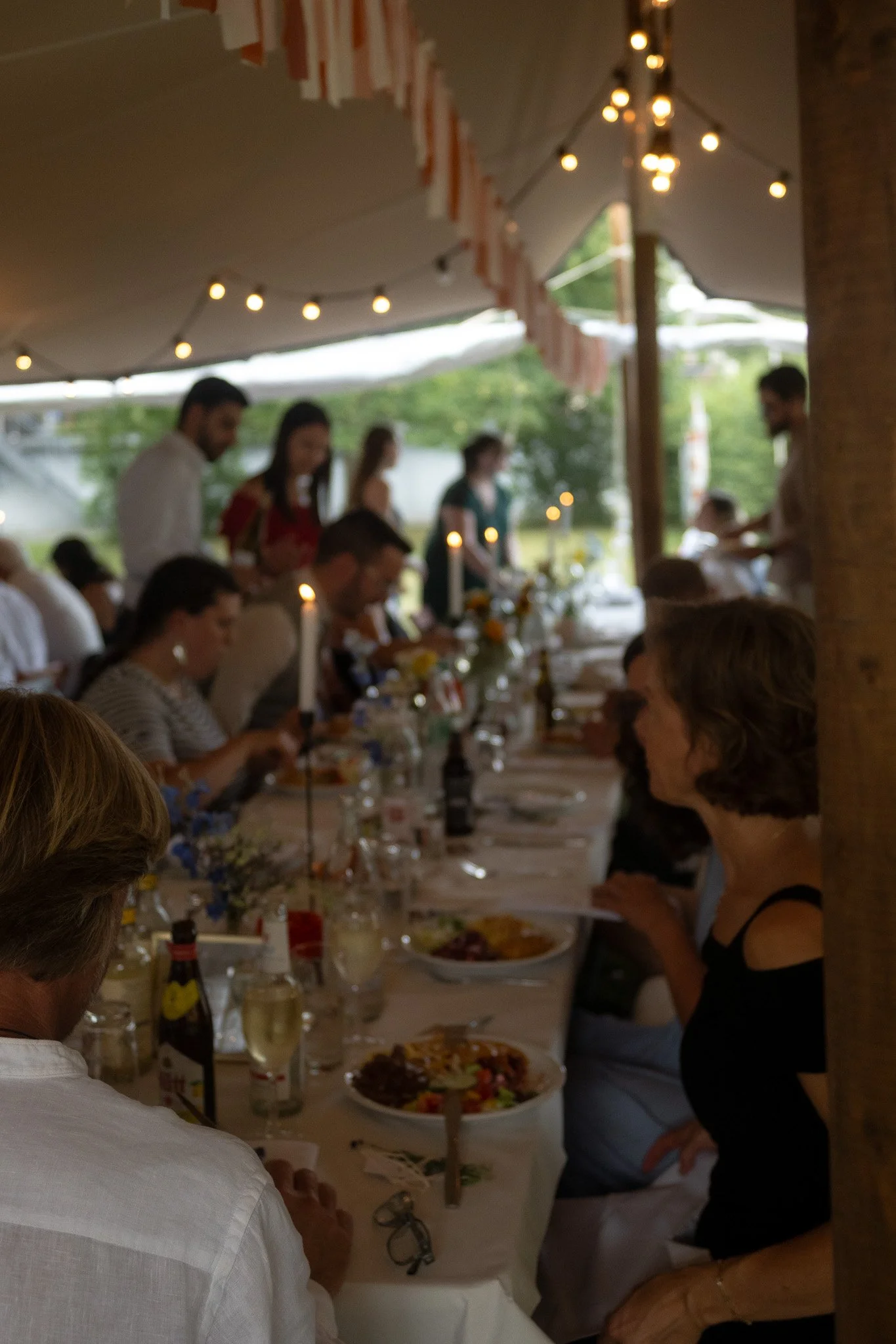 Blick auf eine festliche Dinner-Feier unter einem Pavillon mit mehreren Gästen, die an einem langen Tisch sitzen, Essen genießen und sich unterhalten; Dekoration mit Lichterketten und Blumen. Es ist wahrscheinlich ein Abendessen bei einem besonderen 