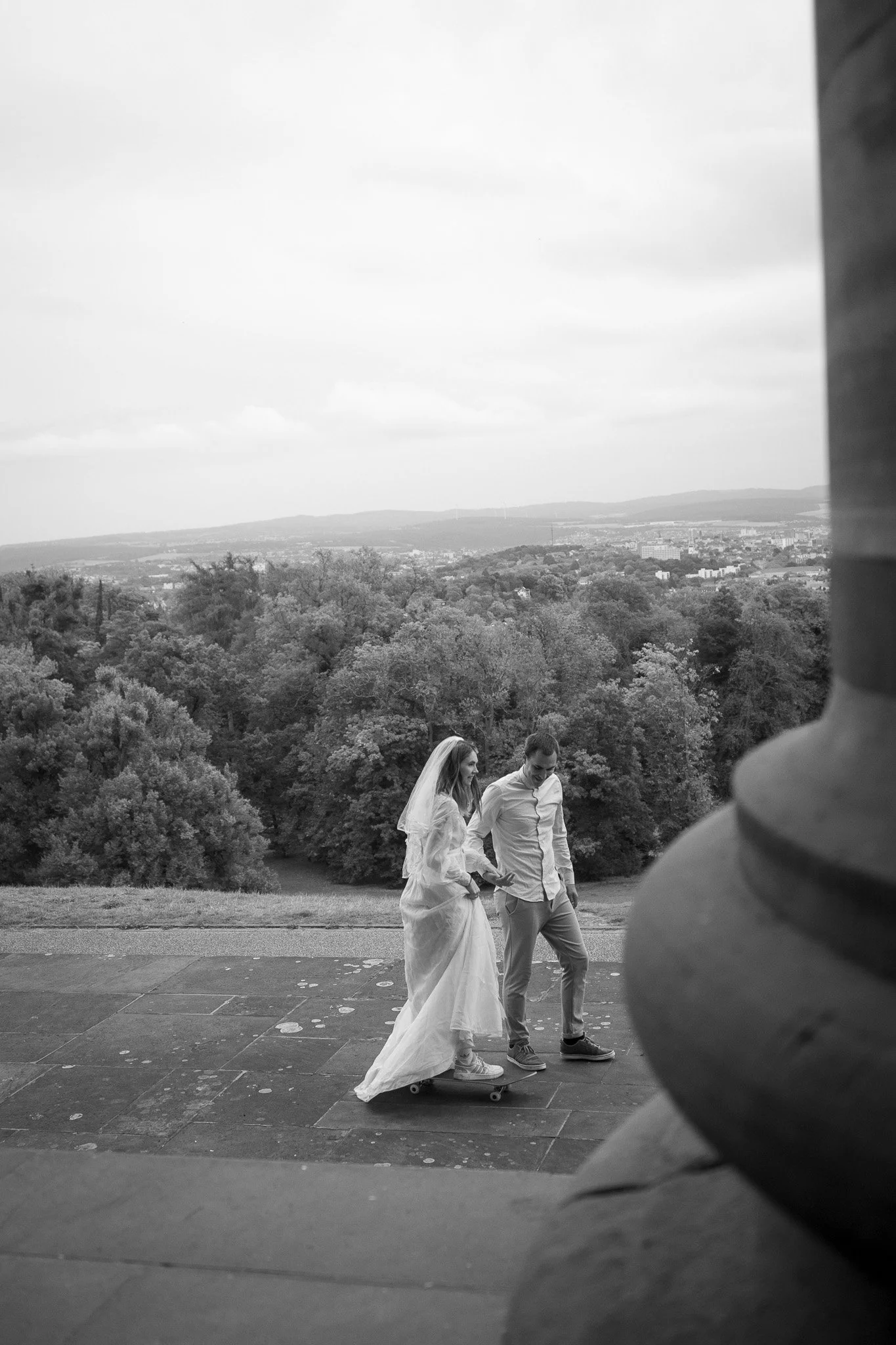 Braut und Bräutigam auf einem Skateboard, draußen auf einer Terrasse mit Blick auf einen bewaldeten Hügel und eine Stadt im Hintergrund, in Schwarz-Weiß.
