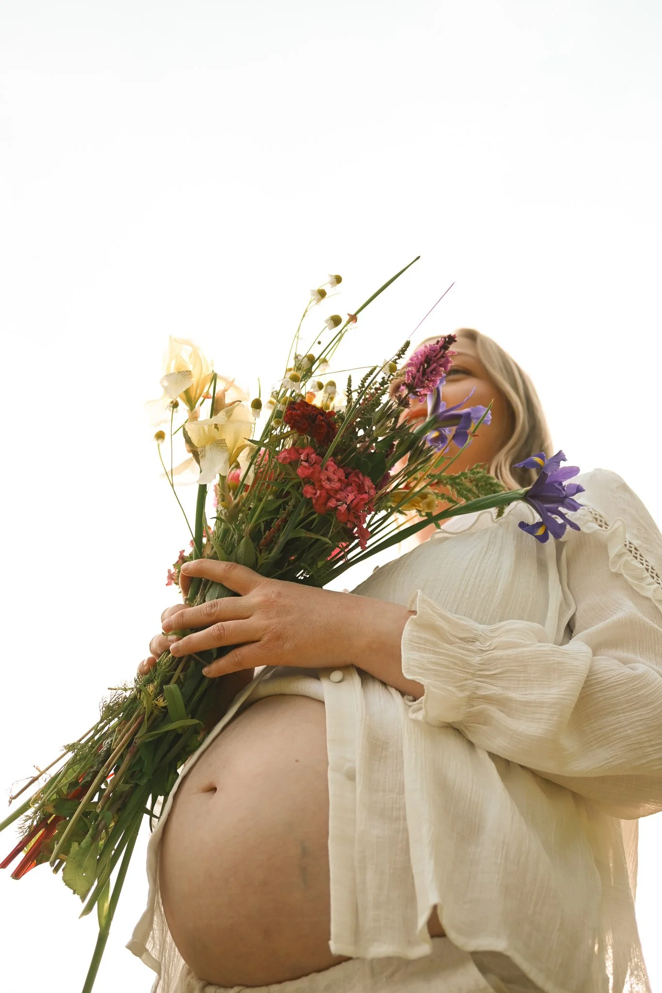 Schwangere Frau mit langem haare und weißen Kleid hält einen bunten Blumenstrauß vor ihrem bauch