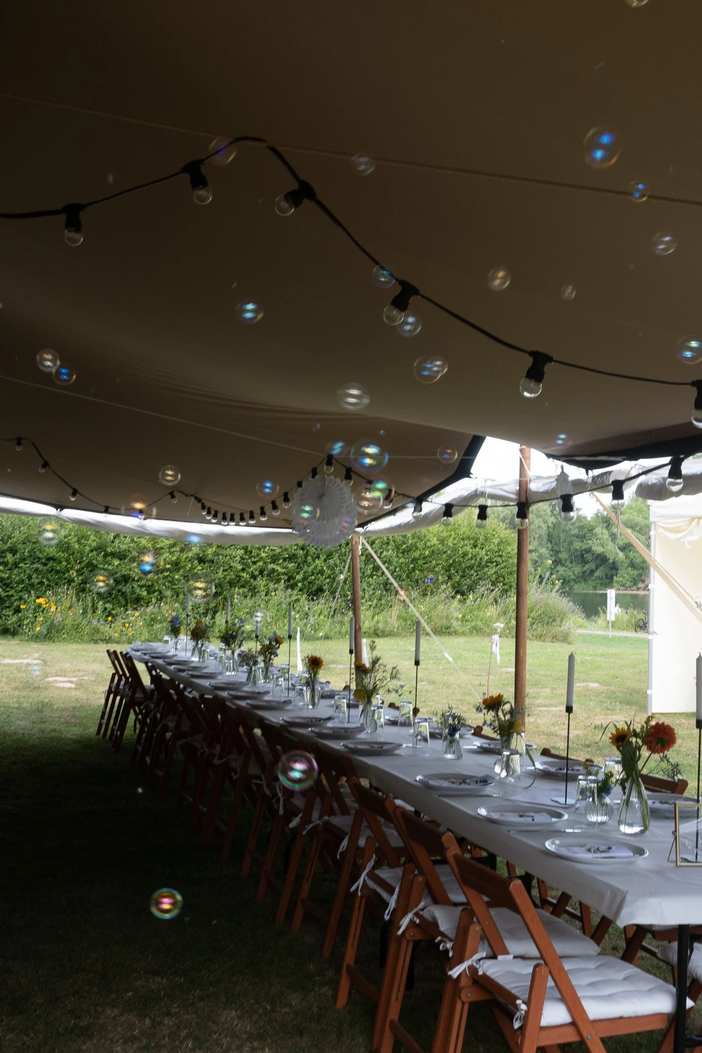 Lange Tafel unter einem großen Zelt, dekoriert mit Sonnenblumen in Vasen und Kerzen, im Freien bei einer Veranstaltung, mit hängen gelassenen Lichterketten und schwebenden Seifenblasen.