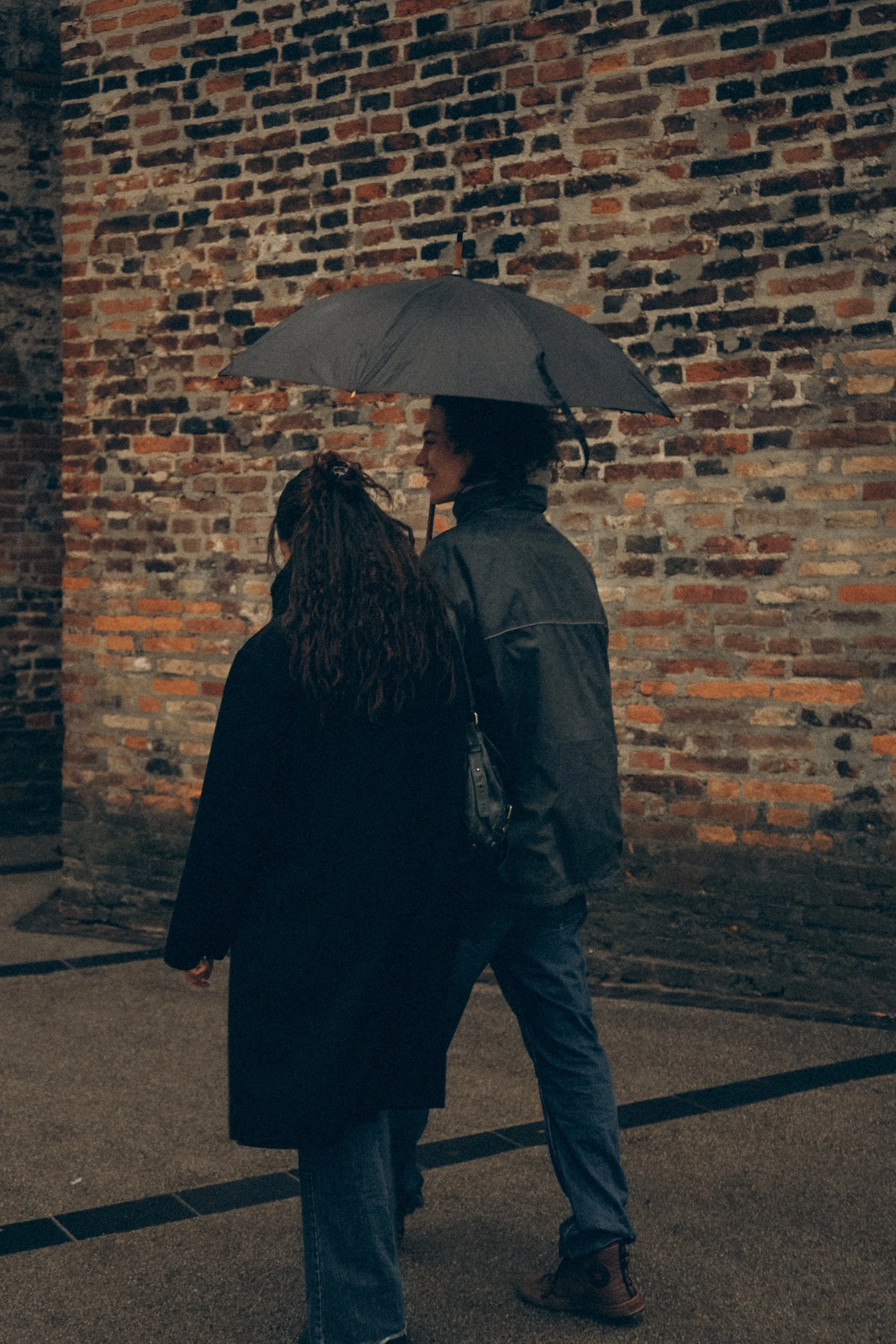 Zwei Frauen unter einem grauen Regenschirm, die sich auf einer regnerischen Straße neben einer Altsteinmauer befinden, im Abendlicht.