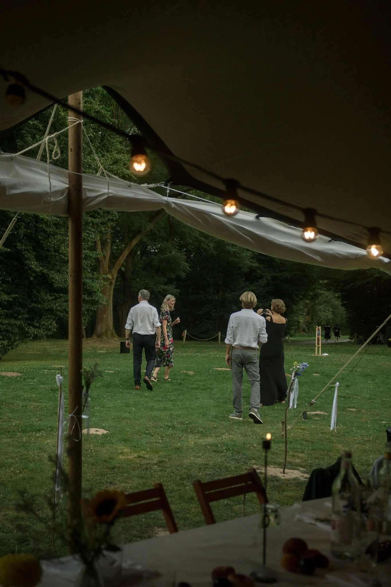 Menschen auf einer Veranstaltung im Freien, zu sehen sind vier Personen, die sich unterhalten oder fotografieren, im Hintergrund sind Bäume und ein Rasenbereich. Die Szene ist abends, beleuchtet von Lichterketten.