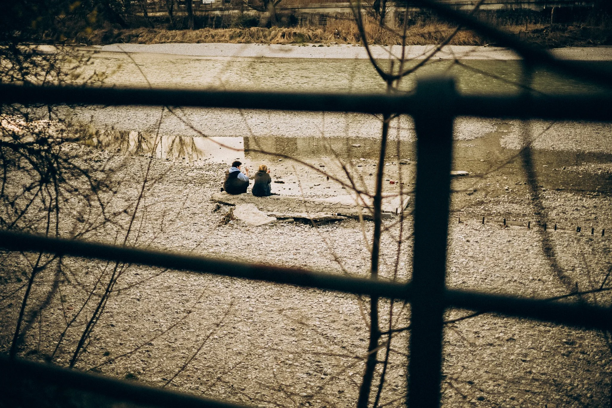 Zwei Menschen sitzen bei Sonnenuntergang am Ufer eines ausgetrockneten Flusses, gesehen durch ein Gitter.