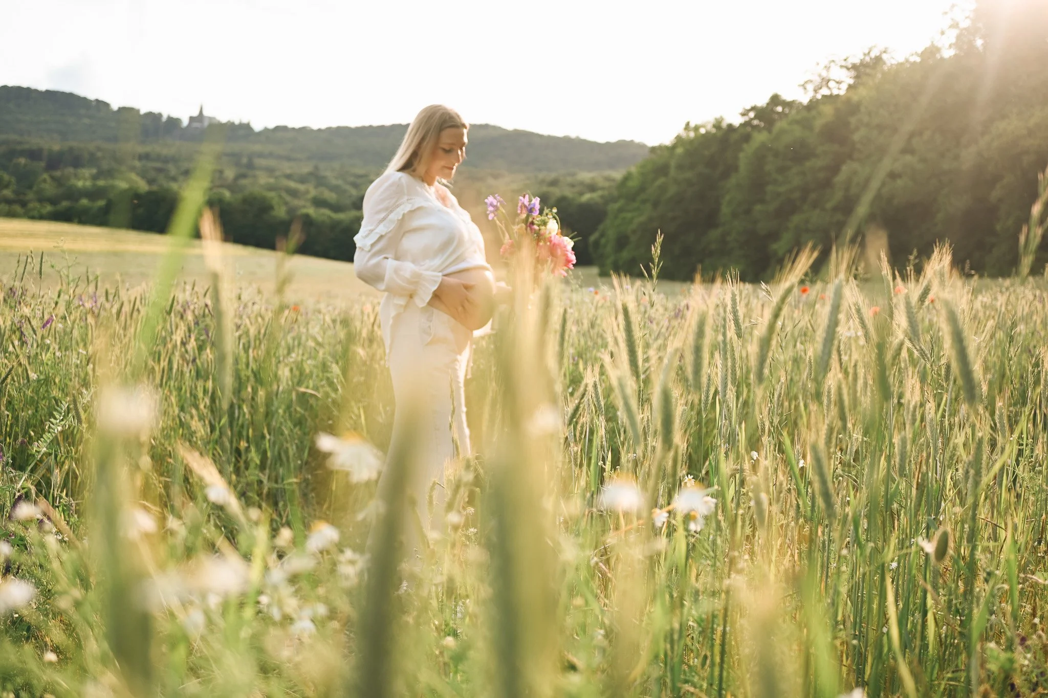 Schwangere Frau in einem weißen Kleid hält einen Blumenstrauß in einem grünen Weizenfeld bei Sonnenuntergang.
