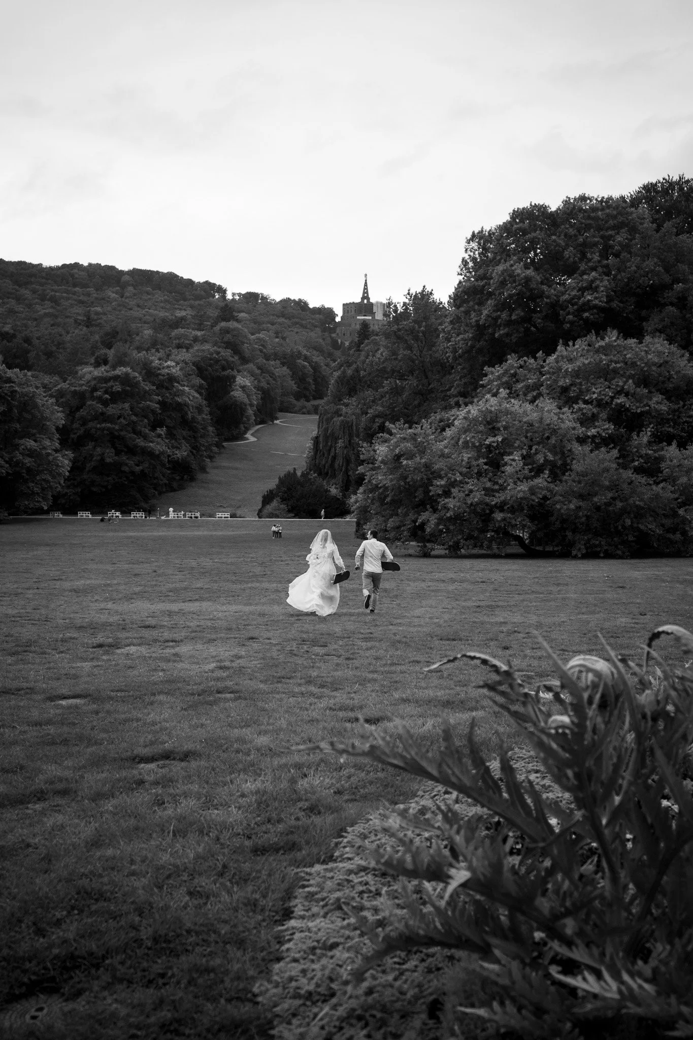 Ein Brautpaar läuft Hand in Hand durch einen großen, offenen Park mit viel Grün und Bäumen, einer im Hintergrund auf einem Hügel stehenden Kirche oder Kapelle, im schwarzen und weißen Fotostil.