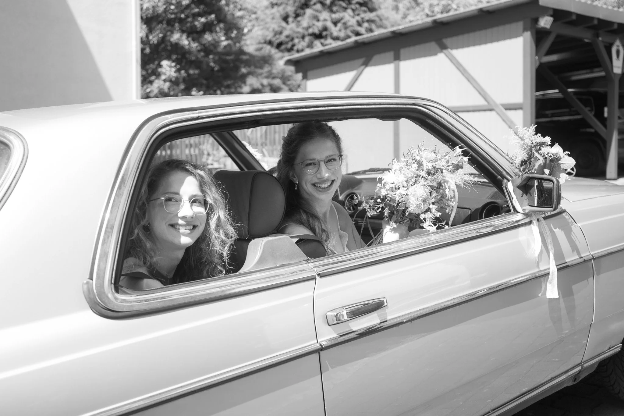 Zwei Frauen fahren in einem Oldtimer-Cabrio, die Sitze sind mit Blumen geschmückt, lachend und glücklich, vermutlich auf dem Weg zu einer Hochzeit.