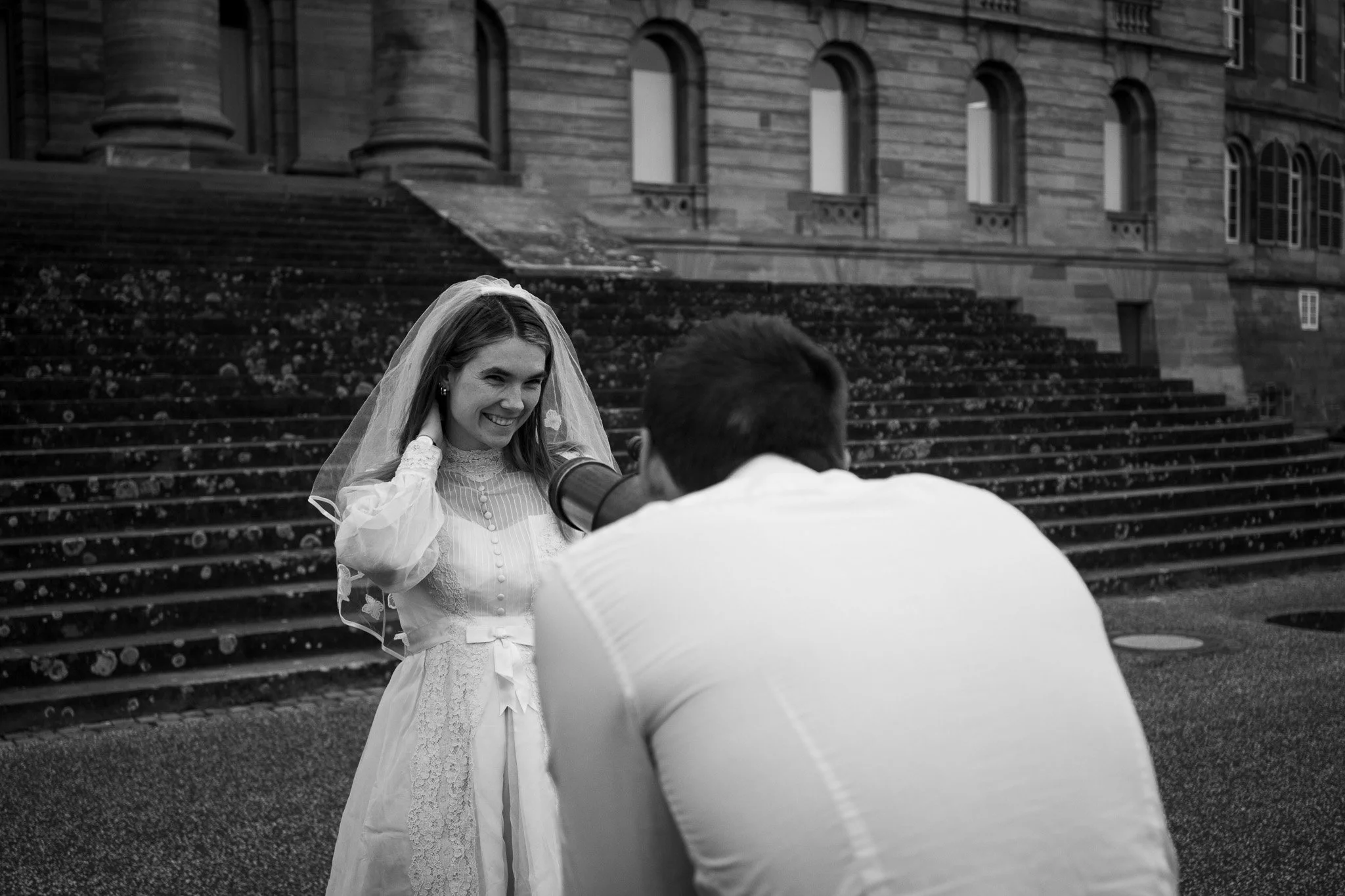 Braut, fotografiert bei einem Fotoshooting vor einem historischen Gebäude, trägt ein Hochzeitskleid mit Schleier, während der Fotograf von hinten fotografiert.
