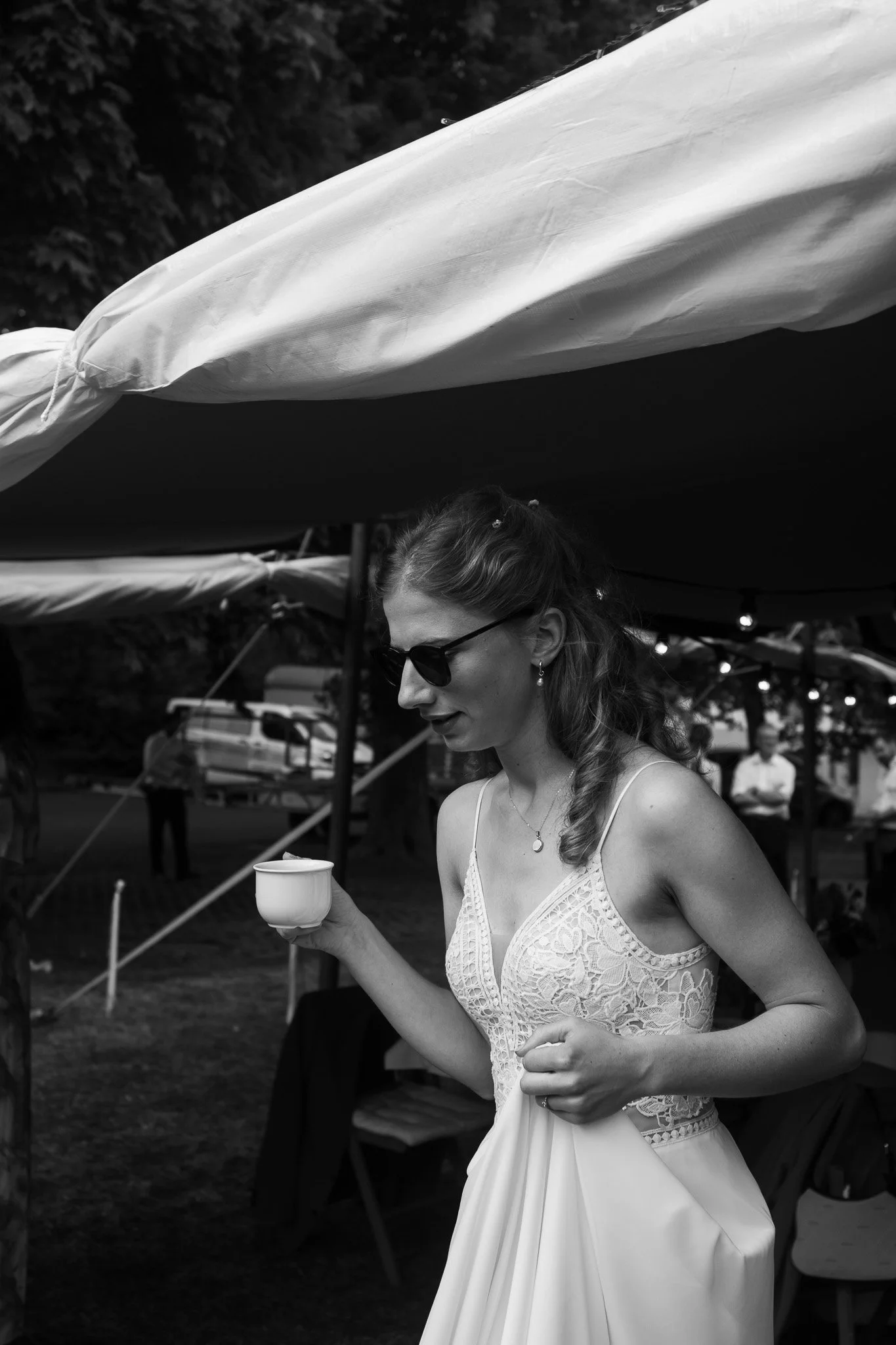 Frau in einem weißen Kleid mit Spaghettiträgern, Sonnenbrille und Ohrringen, die unter einem Pavillon steht, hält eine Tasse in der Hand und schaut nach unten.