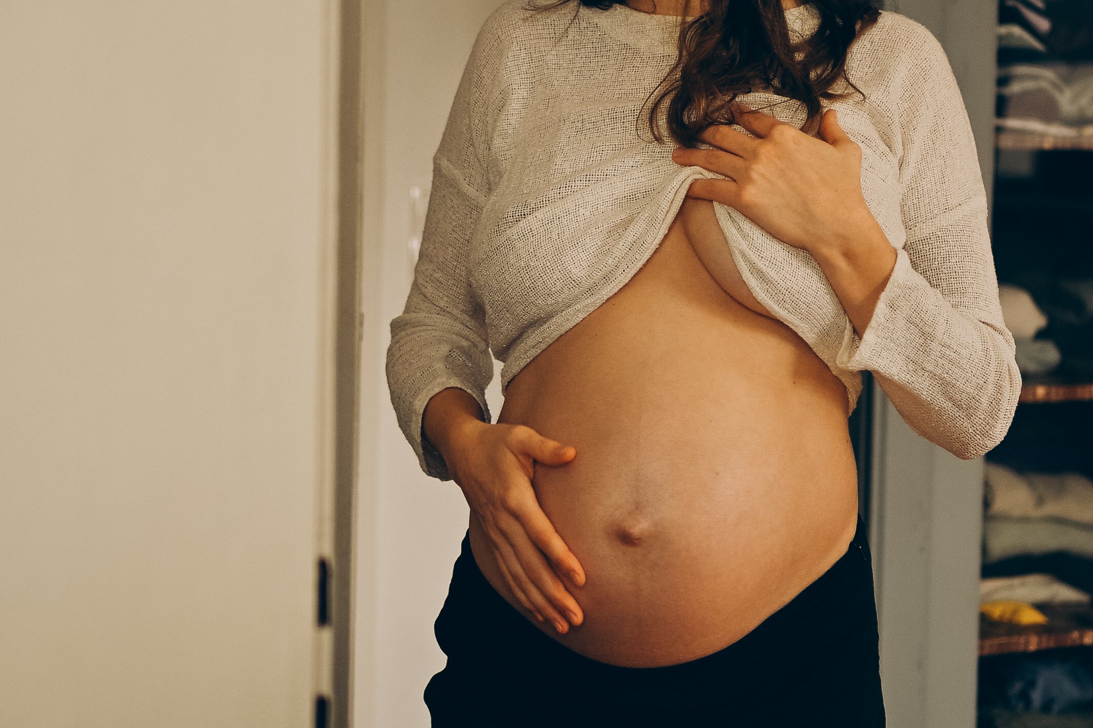 Eine schwangere Frau hebt ihr Hemd hoch, um ihren Babybauch zu zeigen, während sie im Spiegel blickt.