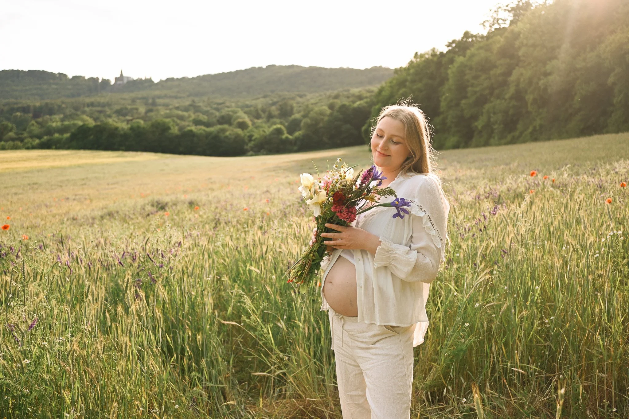 Schwangere Frau in weißer Kleidung mit Blumenstrauß in einem Feld bei Sonnenuntergang.