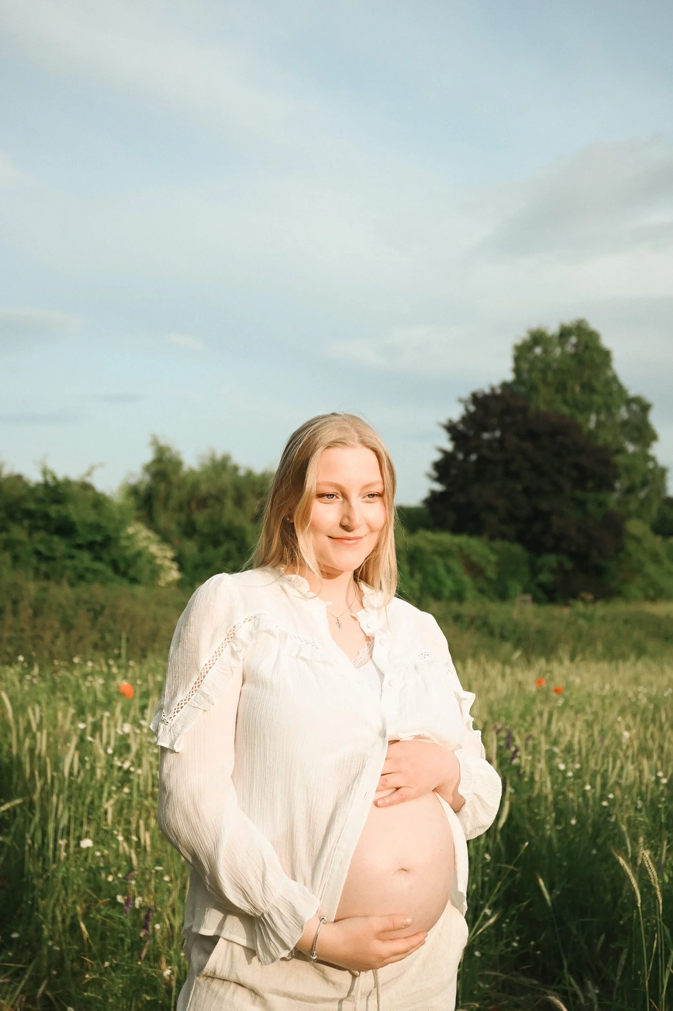 Schwangere Frau im weißen Kleid steht in einem Feld mit grünen Pflanzen und Bäumen, bei Sonnenuntergang, lächelt.