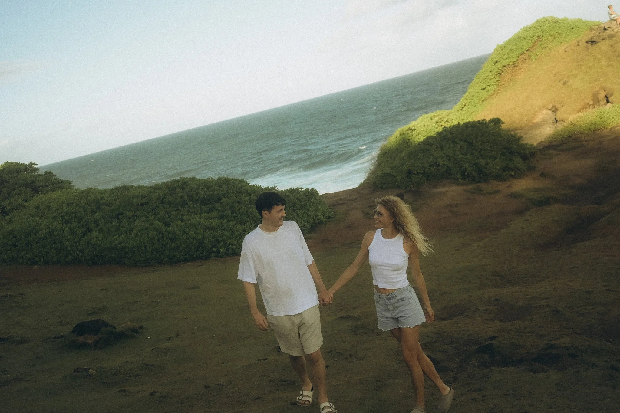 Ein Mann und eine Frau, die sich an einem Strand bei Sonnenuntergang halten, mit Blick aufs Meer und eine grüne Klippe im Hintergrund.