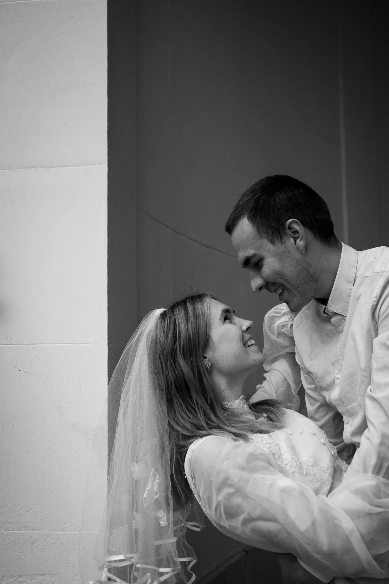 Braut und Bräutigam, die bei ihrer Hochzeit lächelnd und sich in die Augen schauend in einem schwarzen-weißen Foto. Die Braut trägt einen Schleier und ein Hochzeitskleid, der Bräutigam trägt ein Hemd.
