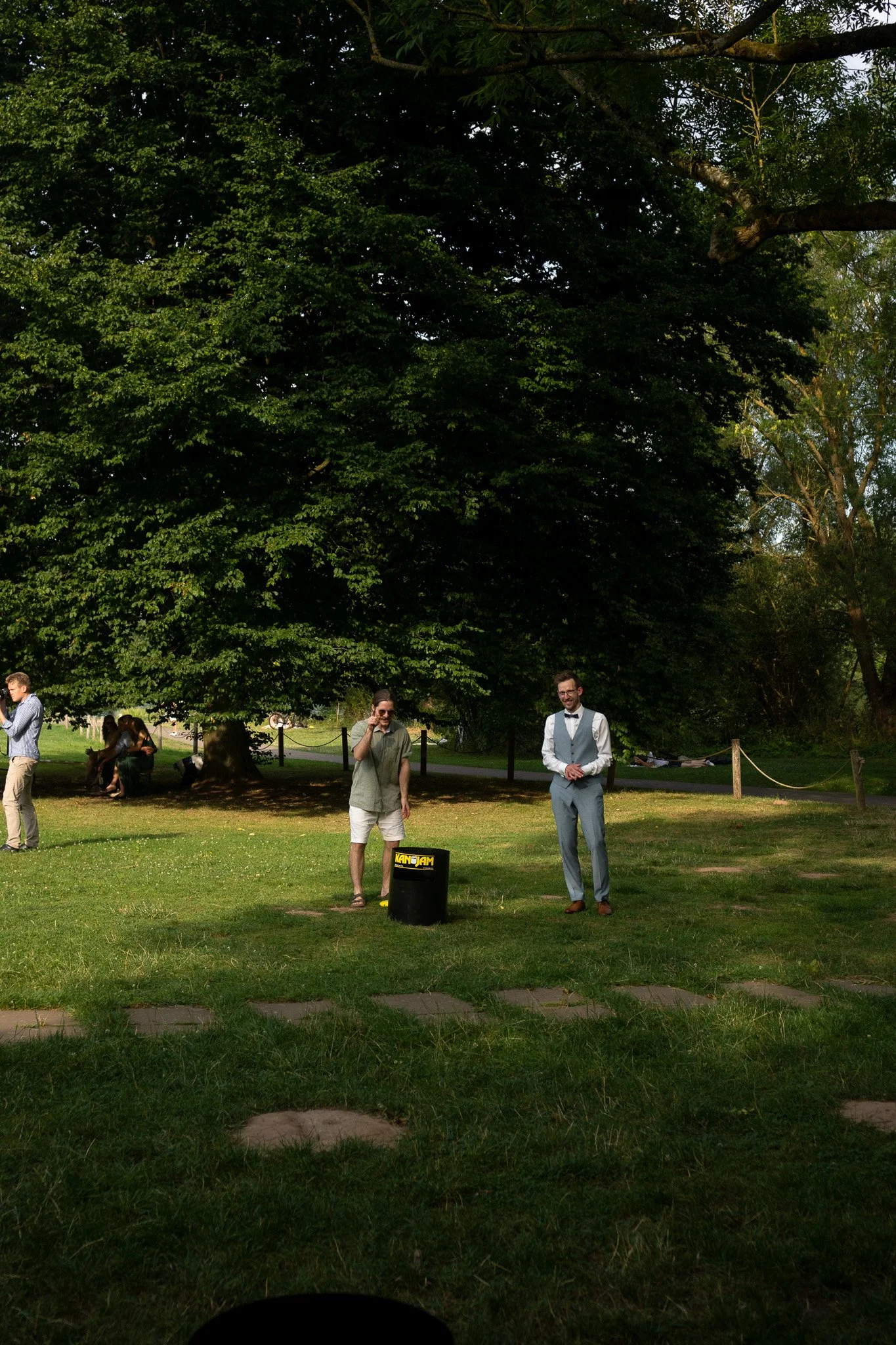 Zwei Männer stehen im Park, einer trägt eine Weste und Krawatte, der andere trägt eine Short und ein T-Shirt. Im Hintergrund sind Bäume und weitere Personen zu sehen.