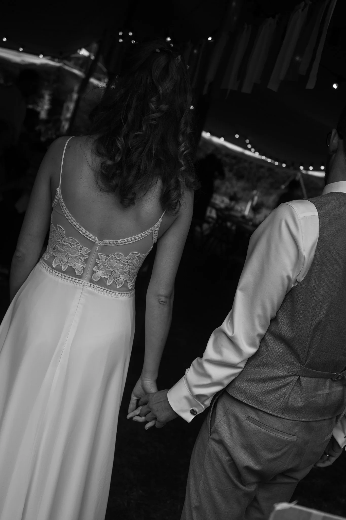 Ein Brautpaar hält Händchen bei einer Hochzeit, die Frau trägt ein weißes Brautkleid mit Spitzeneinsätzen am Rücken, der Mann trägt einen grauen Anzug mit weißem Hemd.