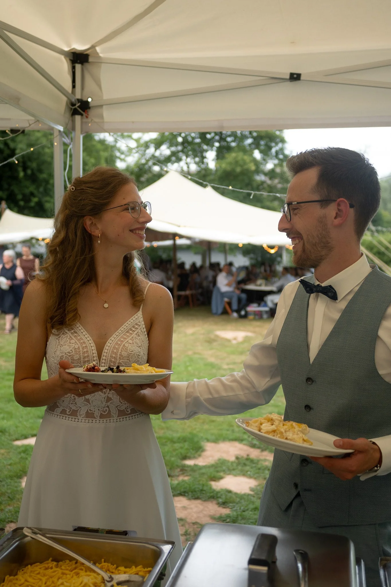 Ein Mann und eine Frau bei einer Hochzeitsfeier unter einem Zelt, die sich gegenseitig lächelnd Essen auf Tellern reichen, im Hintergrund Gäste und Dekorationen.
