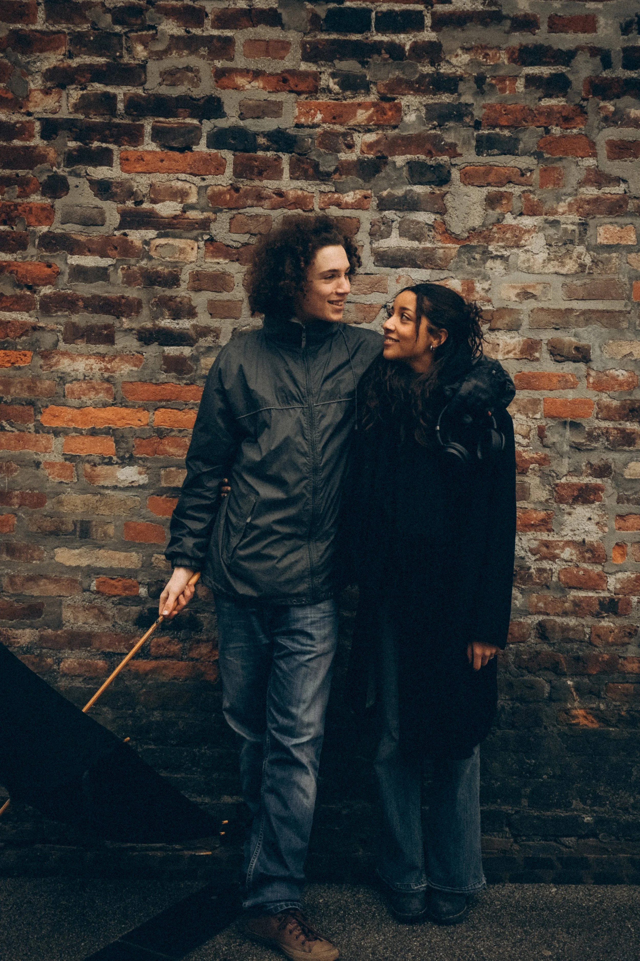 Zwei Menschen, ein Mann und eine Frau, stehen eng an eine Ziegelsteinmauer gelehnt und lächeln sich an.