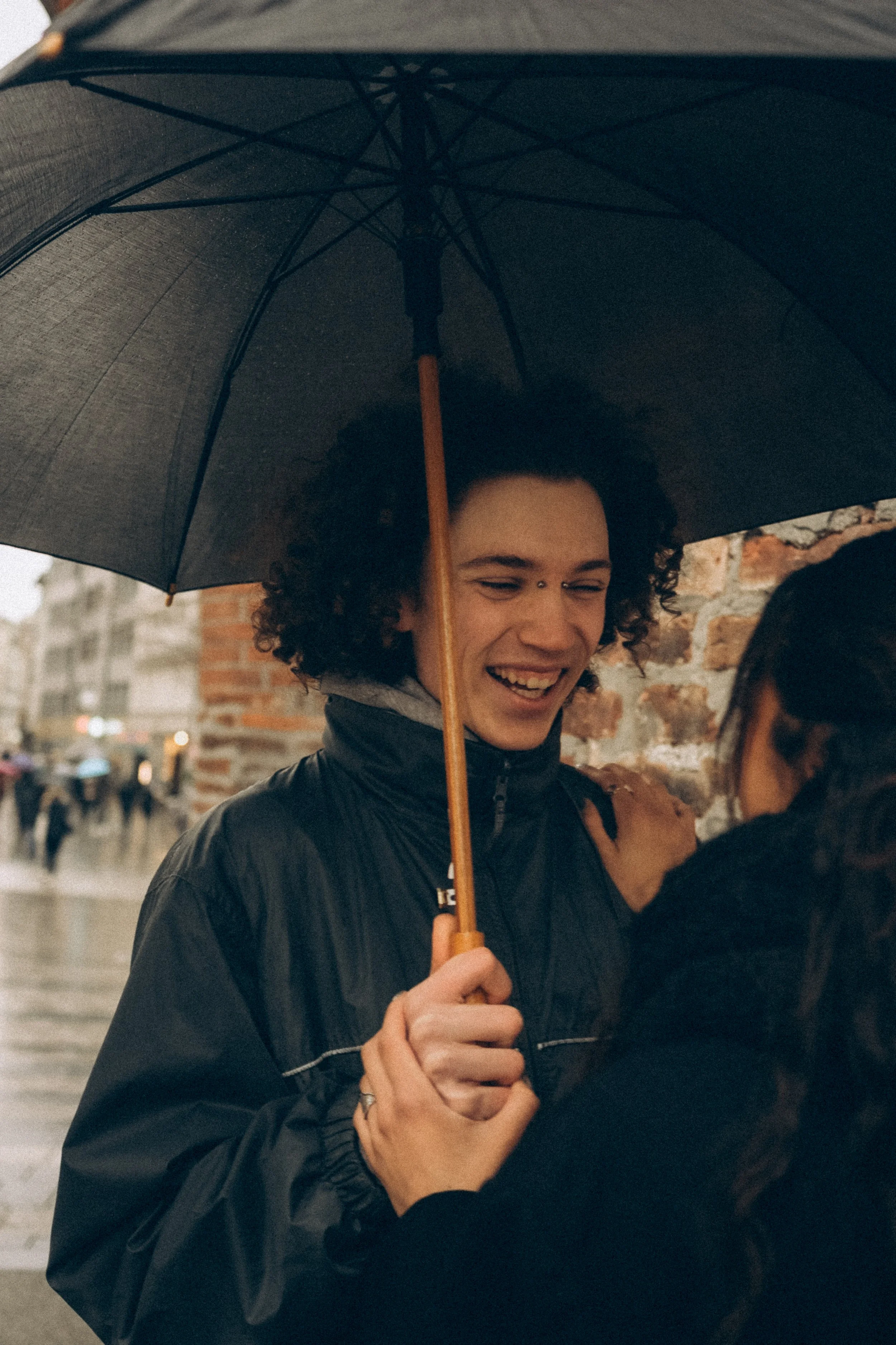 Ein lachendes Paar unter einem schwarzen Regenschirm steht in der Nähe einer Ziegelmauer im Regen, umgeben von einer Stadt mit Fußgängern.
