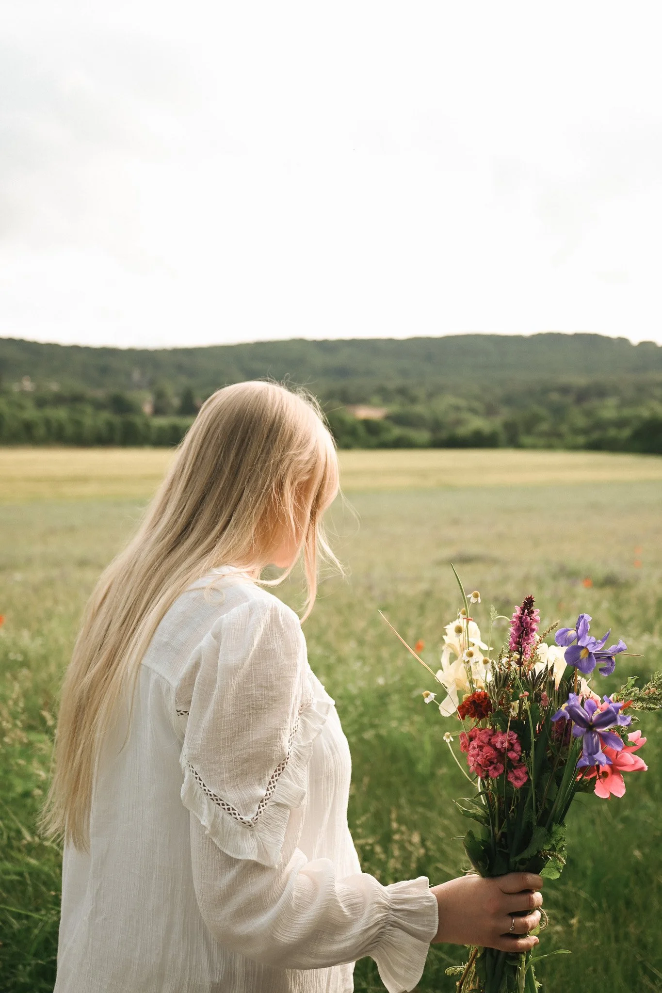 Frau mit langem blondem Haar hält einen Blumenstrauß in einem offenen Feld mit Hügel im Hintergrund, bei bewölktem Himmel.