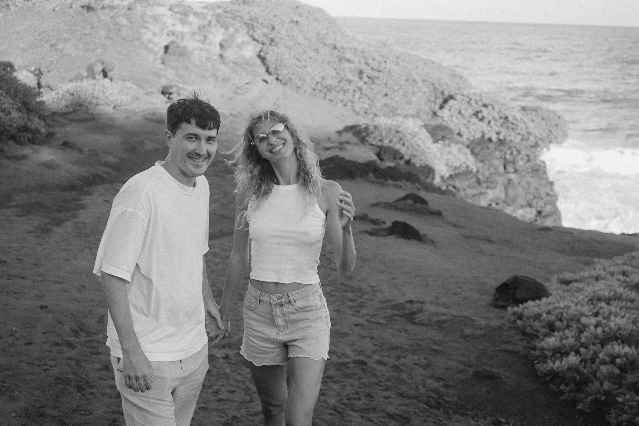 Ein junges Paar steht lachend an einer felsigen Küste mit Wasser im Hintergrund, der Mann trägt ein weißes T-Shirt, die Frau trägt ein weißes Top und Shorts, beide wirken fröhlich und entspannt.