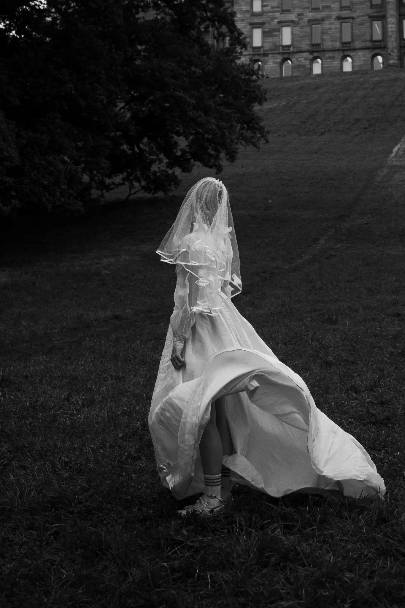Eine Frau in einem weißen Hochzeitskleid mit Schleier steht auf einer Wiese in einem Park, im Hintergrund sind Bäume und ein großes altes Gebäude zu sehen.