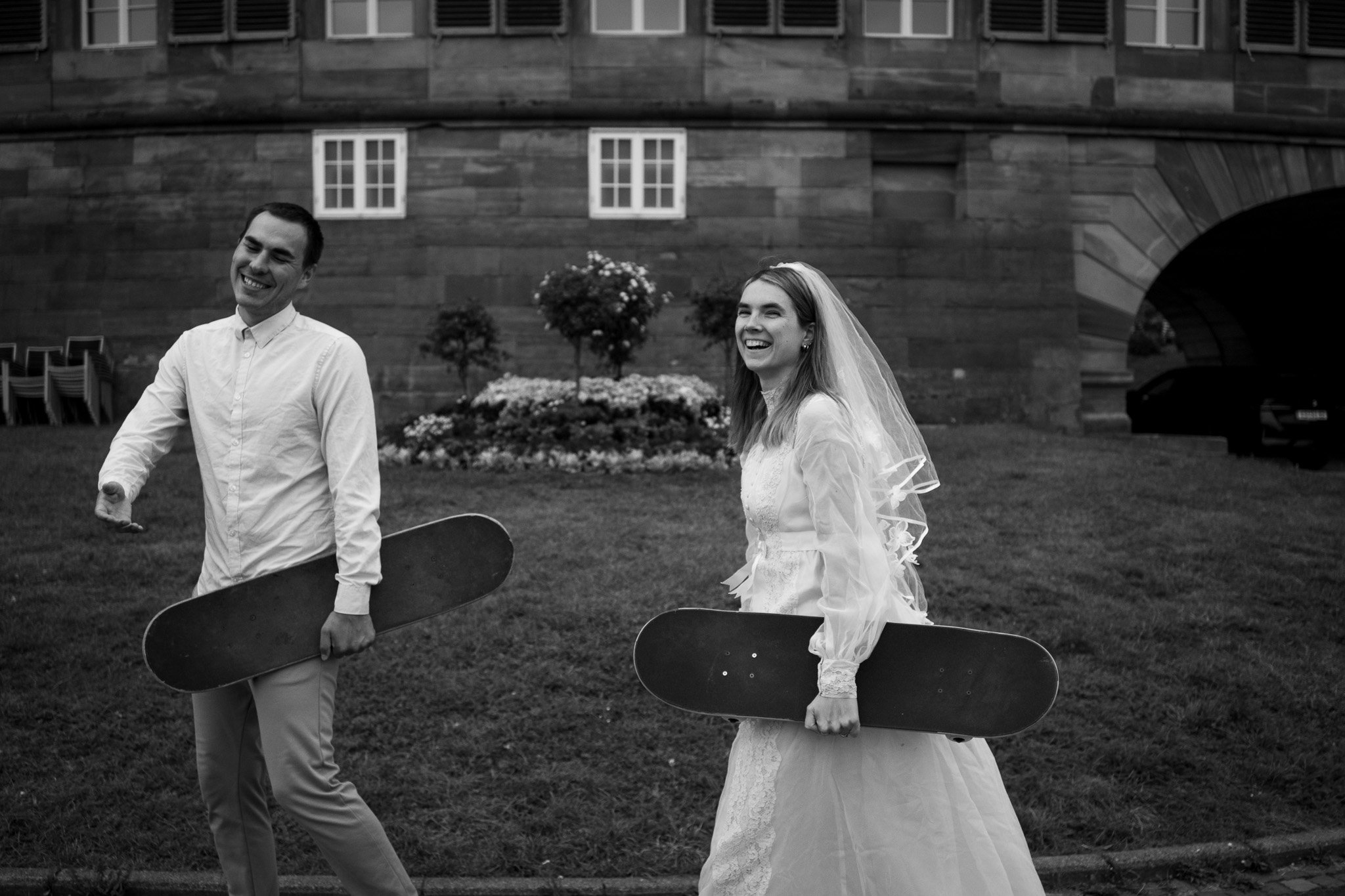 Ein Lächelndes Brautpaar in Hochzeitskleidung, das auf einem Rasen mit Skateboards in der Hand steht, im Hintergrund ein altes Gebäude mit Bäumen und Blumen.