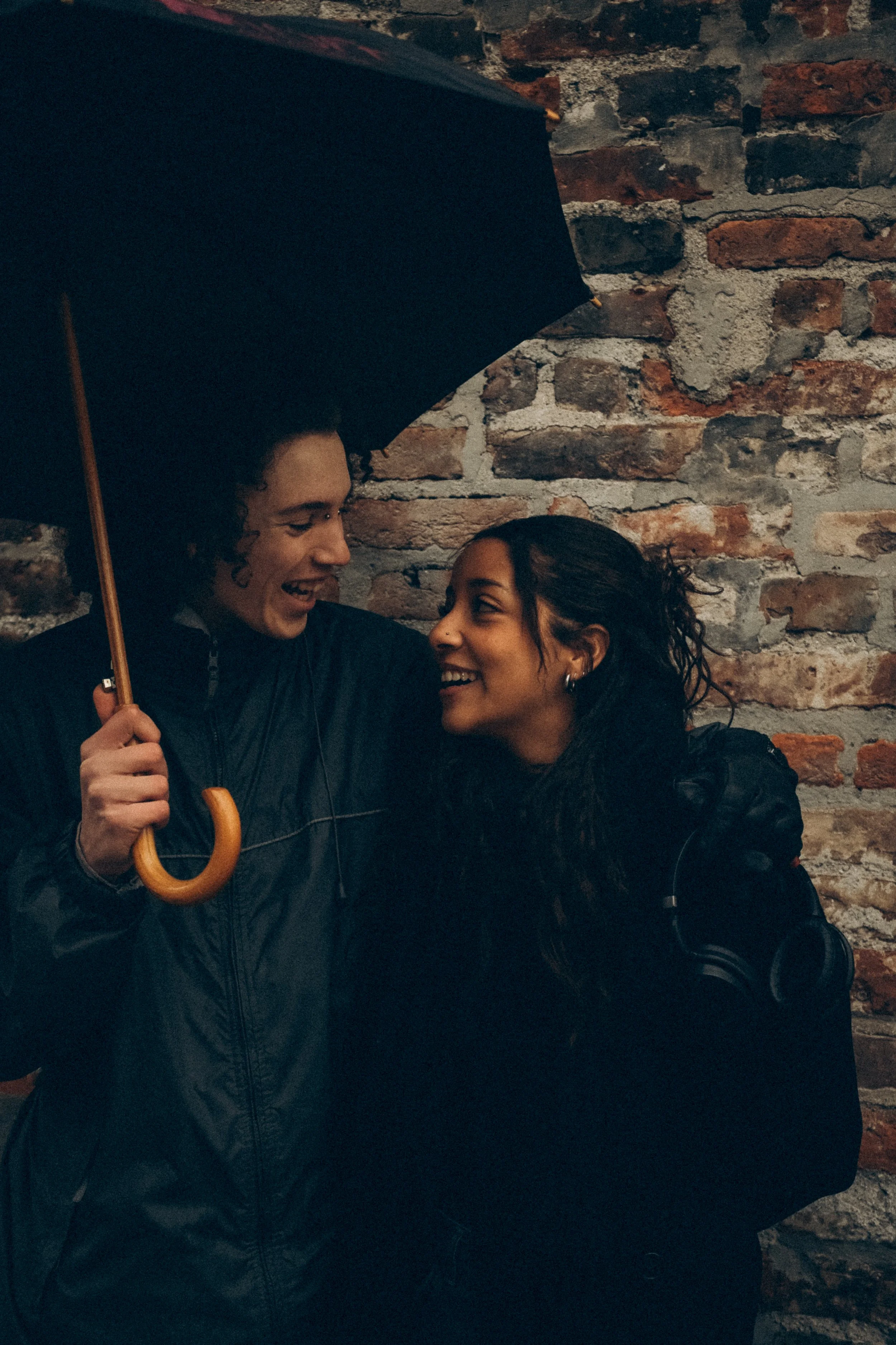 Zwei Menschen unter einem schwarzen Regenschirm, die lachen und sich anlächeln, vor einer alten Ziegelmauer