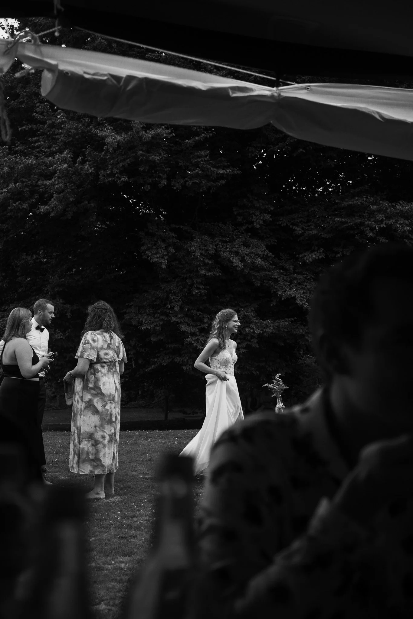Hochzeit im Freien, Braut in Weiß mit langem Kleid, Gäste im Hintergrund, grüne Bäume, schwarze und weiße Fotografie.