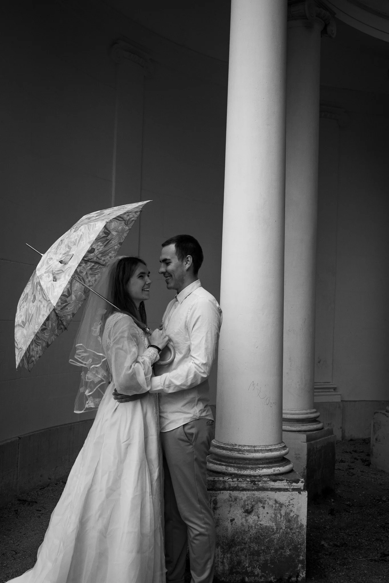 Ein junges Paar in formeller Kleidung, das unter einem Regenschirm lächelnd zusammensteht, neben einer großen Säule in einer Gebäudearchitektur, im Schwarz-Weiß-Format.