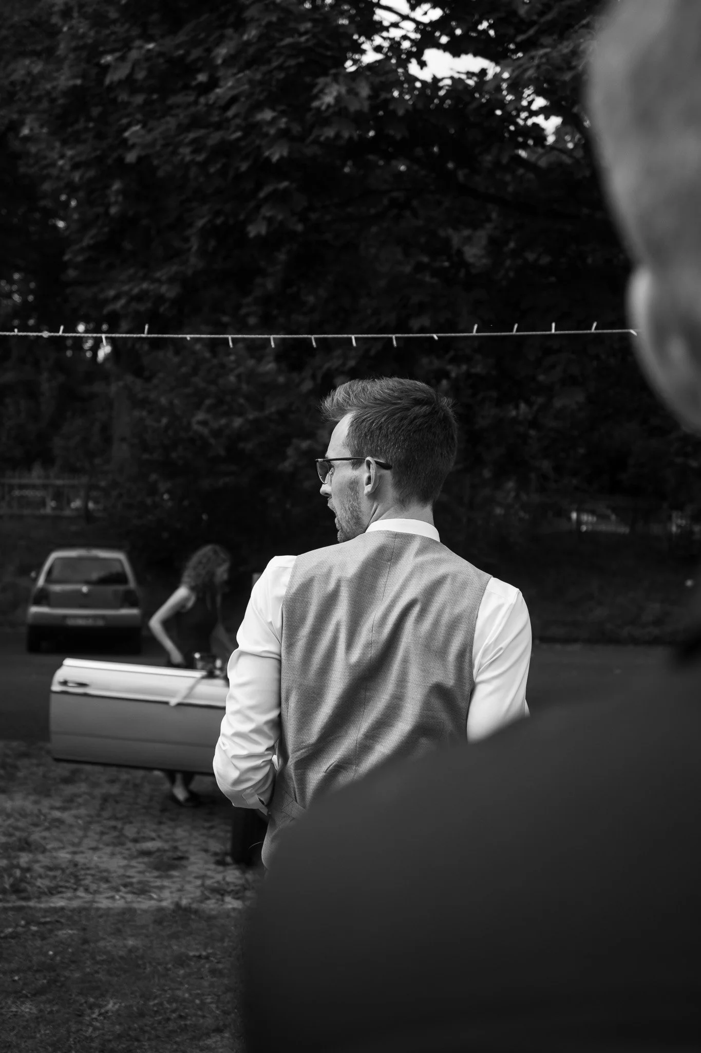 Schwarz-weiß-Foto eines Mannes in einem Anzug, der nach hinten schaut, während eine Frau im Hintergrund mit einem Boot arbeitet.