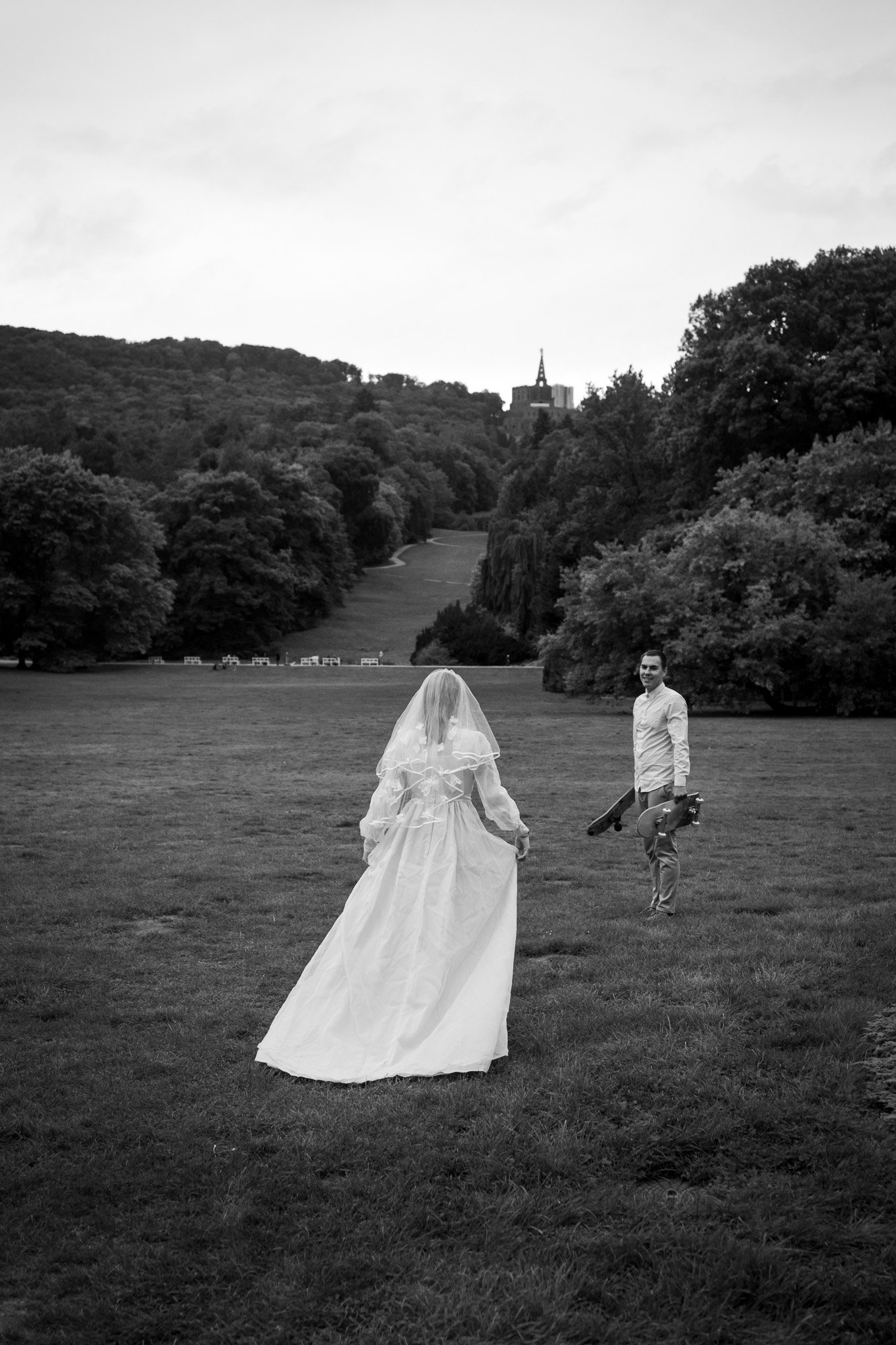 Eine schwarz-weiße Fotografie zeigt eine Frau in einem Brautkleid und Schleier, die auf einer großen Wiese steht, während ein Mann mit einem Skateboard in der Hand Richtung sie blickt, umgeben von Bäumen und Hügeln im Hintergrund.