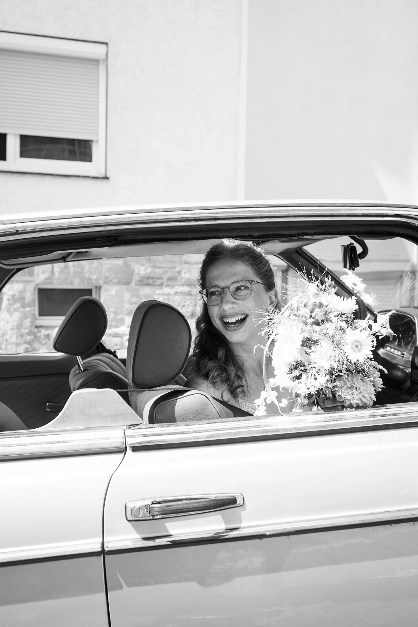 Eine glückliche Frau mit Brille sitzt in einem Auto und hält einen Blumenstrauß in der Hand.