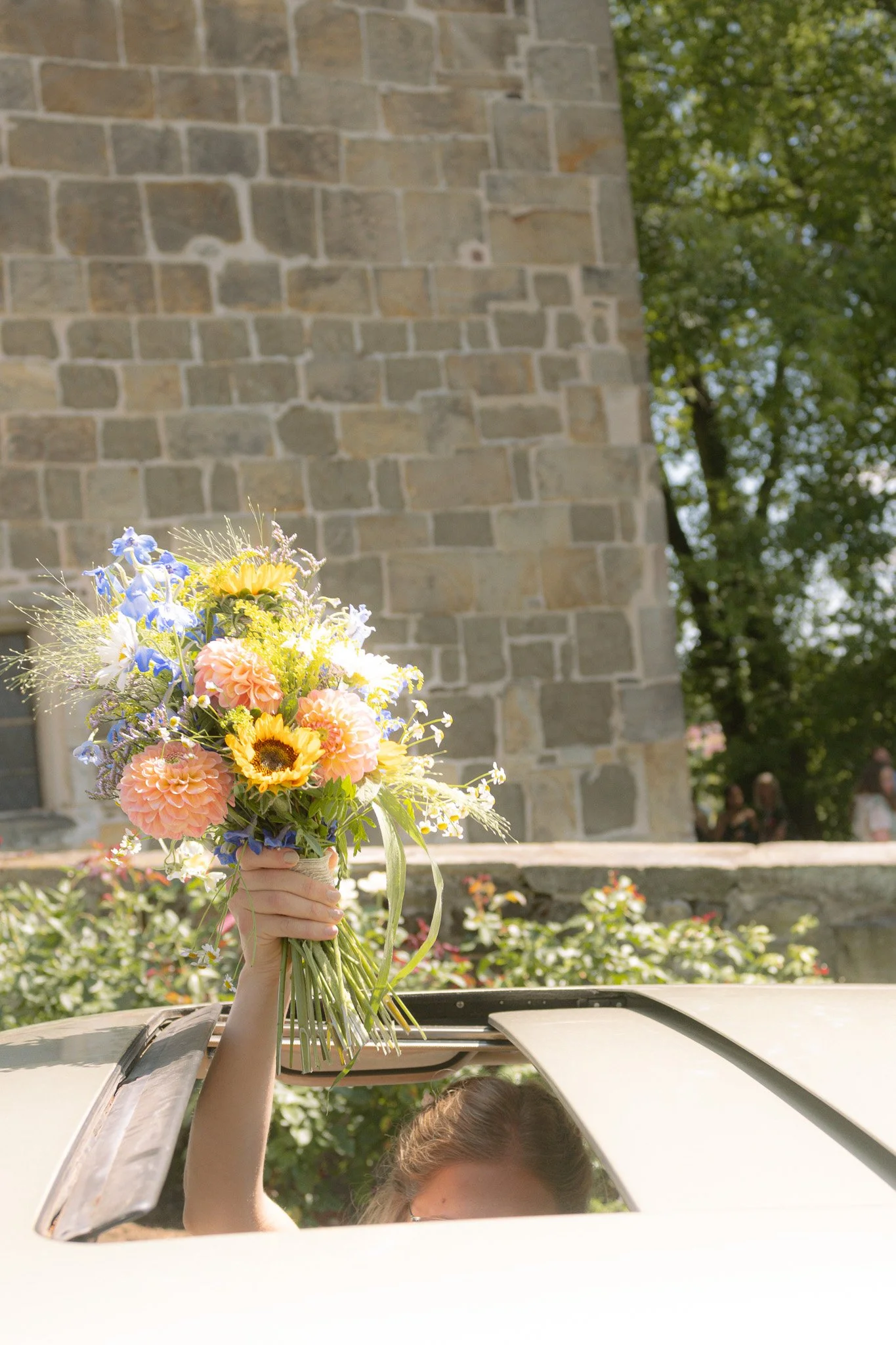 Hand hält bunte Blumenstrauß aus Dahlien, Sonnenblumen, Veilchen und Gräsern aus dem Autofenster heraus.