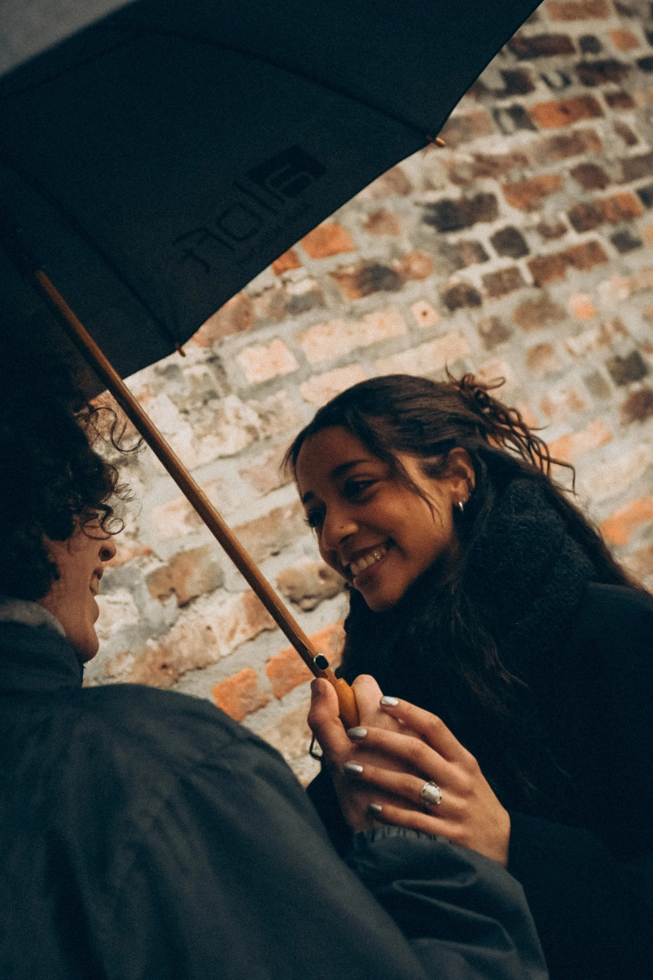 Zwei Menschen lachen und teilen einen Moment unter einem dunkelblauen Regenschirm mit einem Backsteinmauer im Hintergrund.