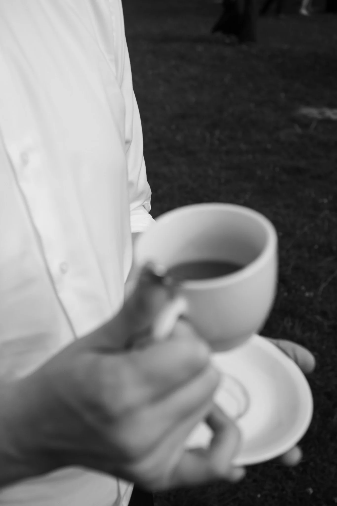Ein Mensch in weißem Hemd hält eine Tasse Kaffee in der Hand, Blick auf die Tasse, Hintergrund unscharf, schwarz-weiß.