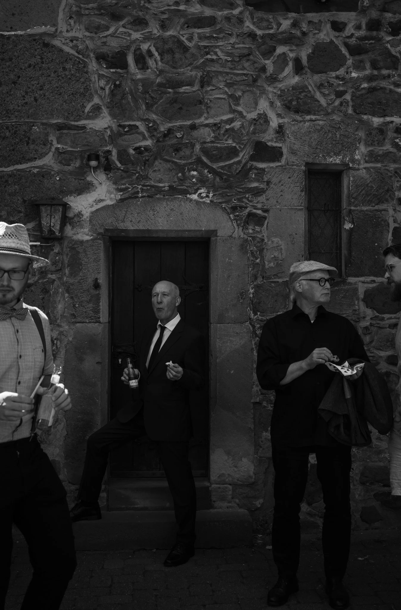 Dreiergruppe von älteren Männern, die an einer alten Steinmauer im Freien stehen, einige essen und trinken, einer im Anzug lehnt an einer Türöffnung, während die anderen beiden in Alltagskleidung stehen und essen.