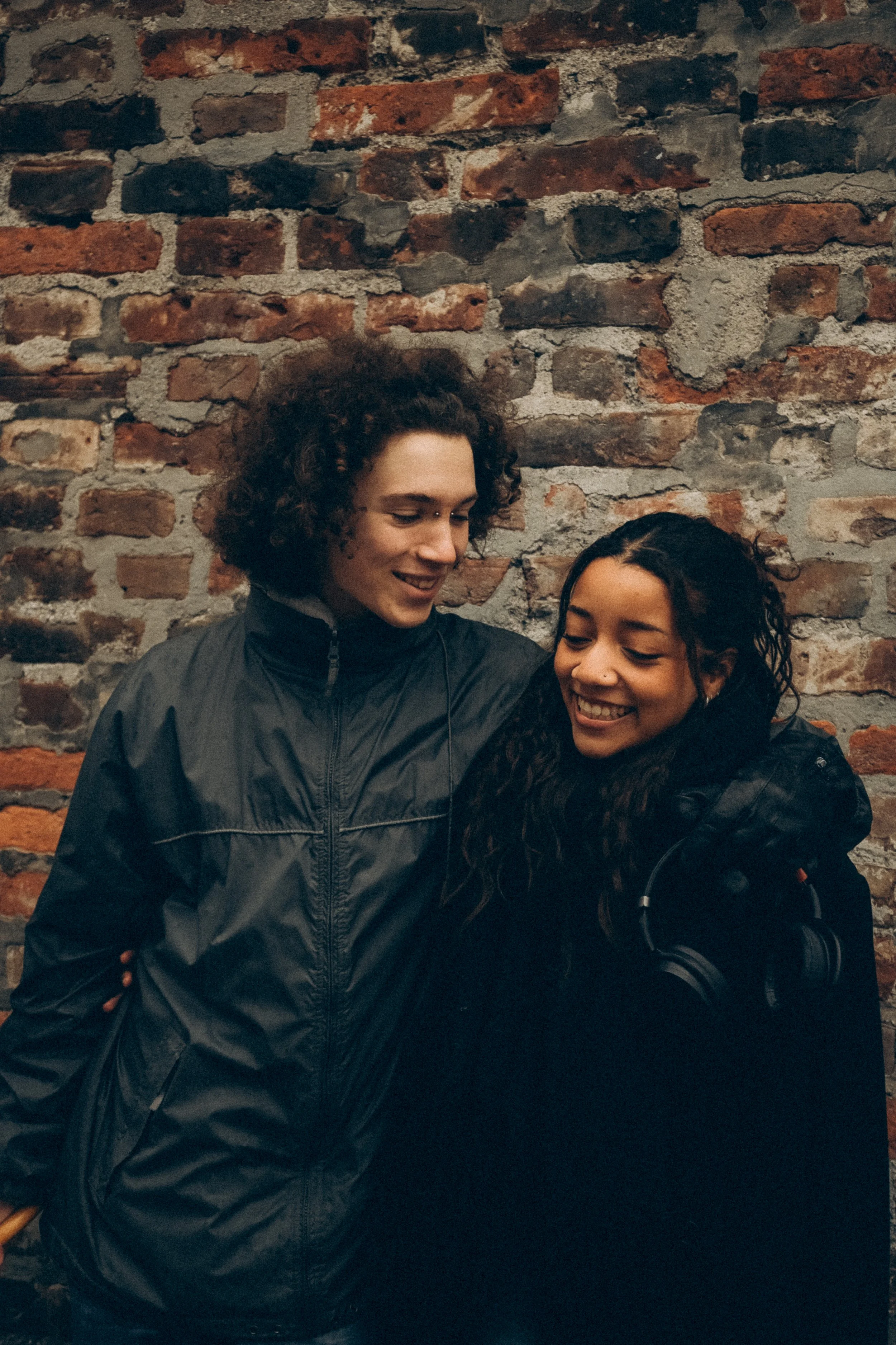Zwei lachende junge Menschen, ein Mann mit lockigem Haar und eine Frau mit längeren, lockigen Haaren, stehen vor einer Backsteinwand. Sie tragen dunkle Jacken und wirken glücklich.