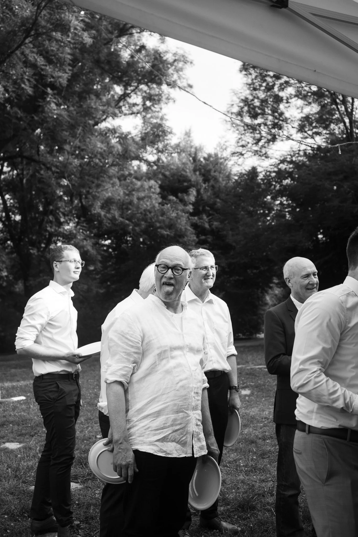 Gruppe von Menschen im Freien, die zu einer Feier oder Veranstaltung vorbereitet sind, einige halten Teller, bei schwarz-weiß Foto, im Hintergrund Bäume.