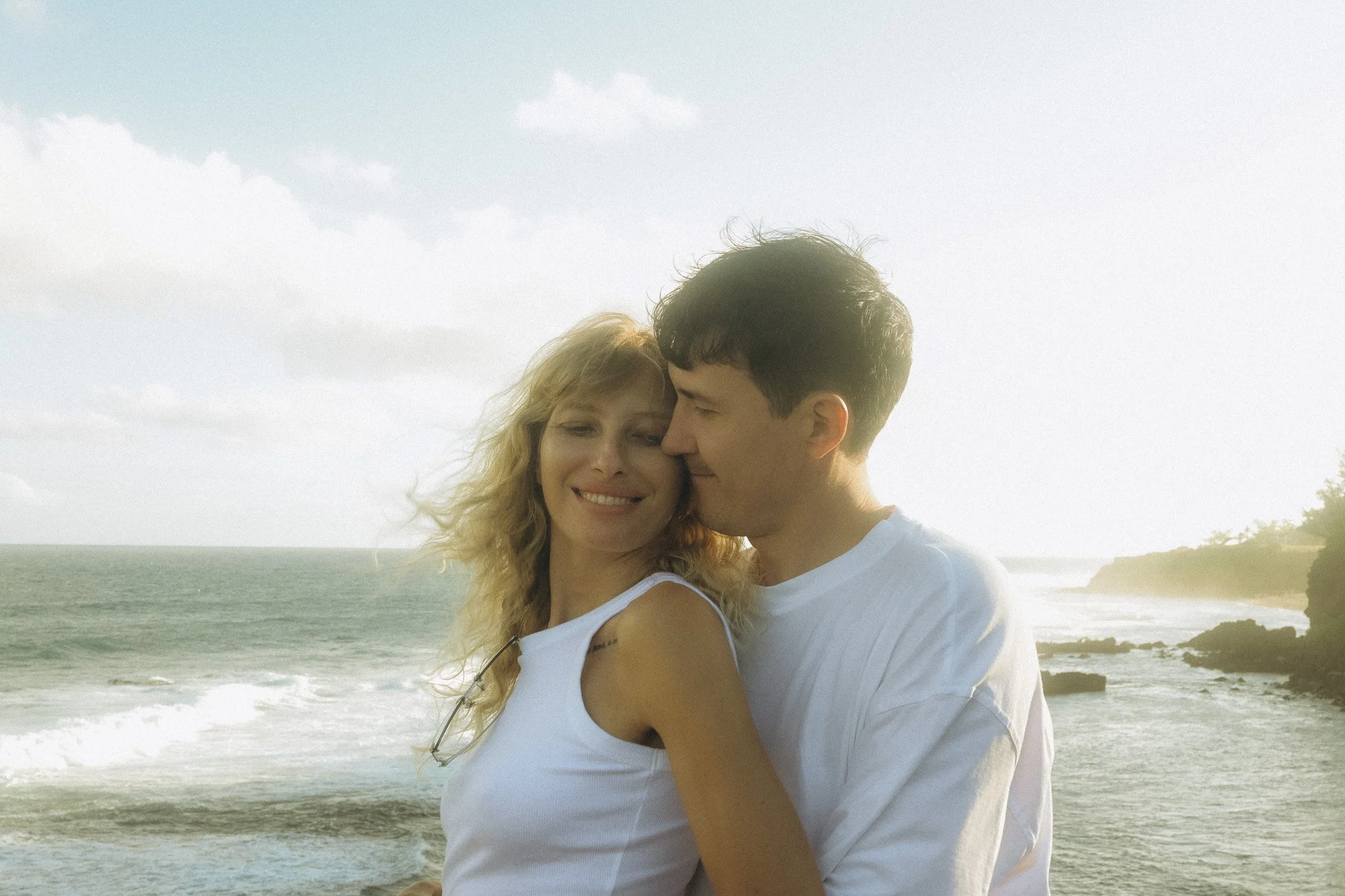 Ein lachendes Paar am Strand bei Sonnenuntergang, die Frau hat blonde, lockige Haare, der Mann hat dunkle, kurze Haare.