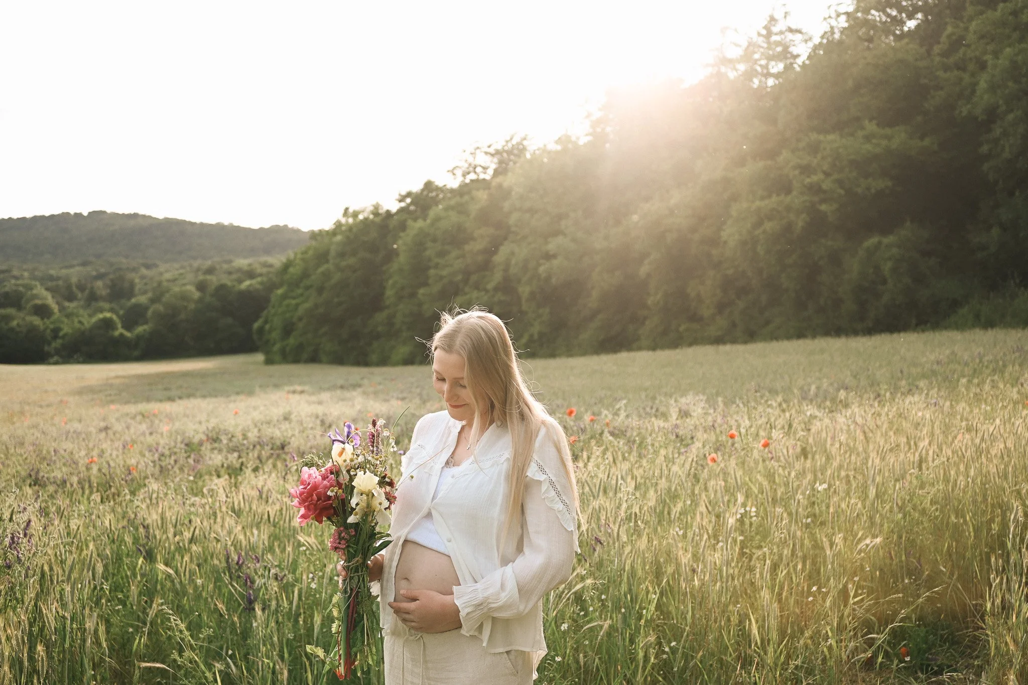 Eine schwangere Frau mit langem blonden Haar trägt ein weißes Kleid und hält einen Blumenstrauß, während sie in einer Wiese bei Sonnenuntergang steht.