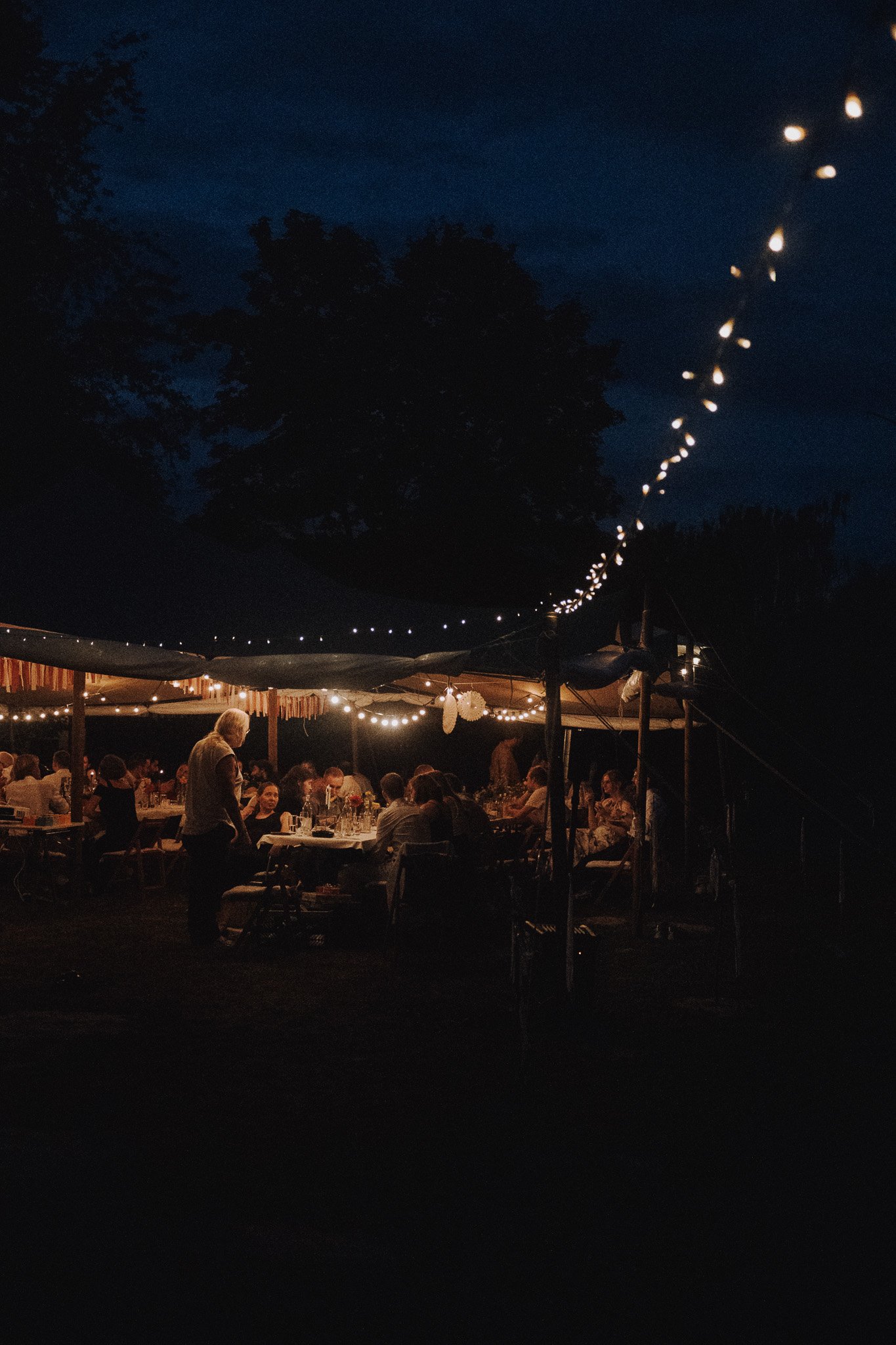 Menschen sitzen bei einer abendlichen Gartenparty unter Lichterketten, die im Dunkeln leuchten.