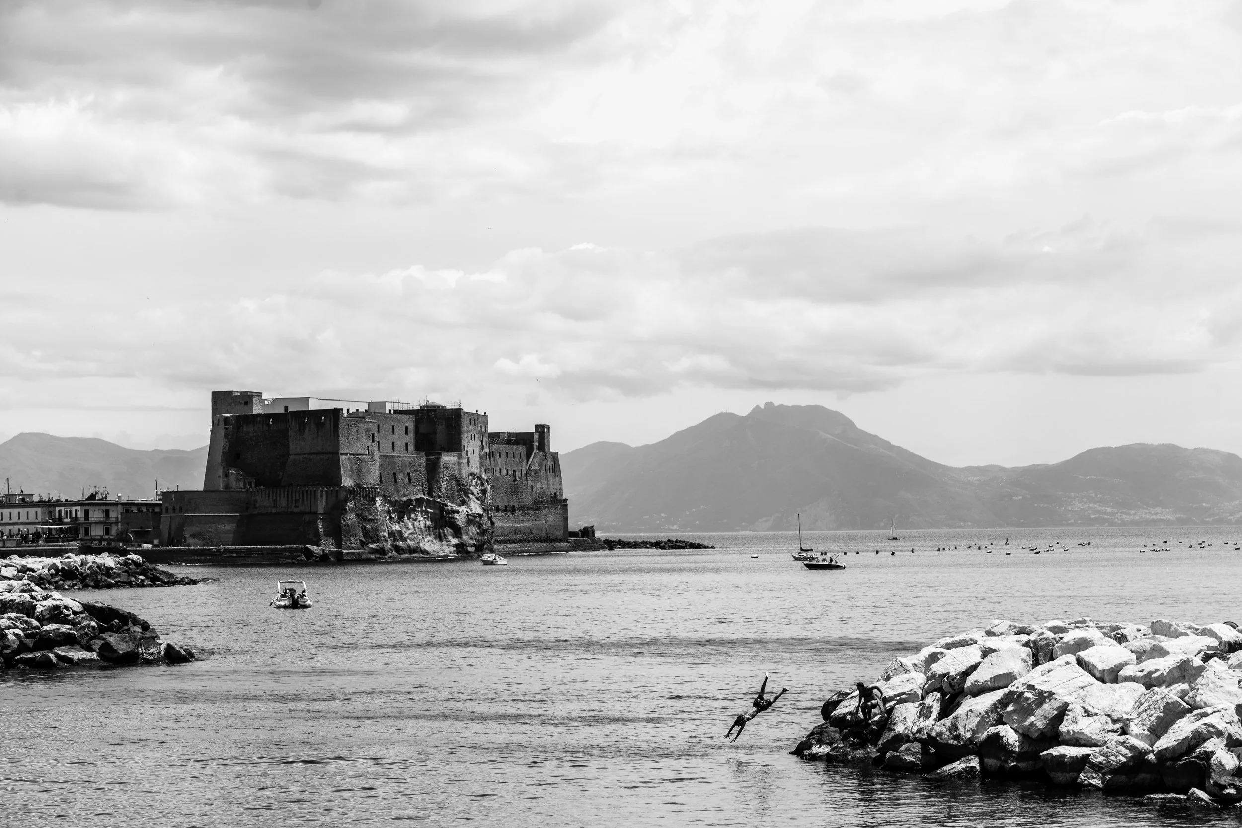A black and white photo of a castle on the shoreline with mountains in the background, boats on the water, and rocks in the foreground.