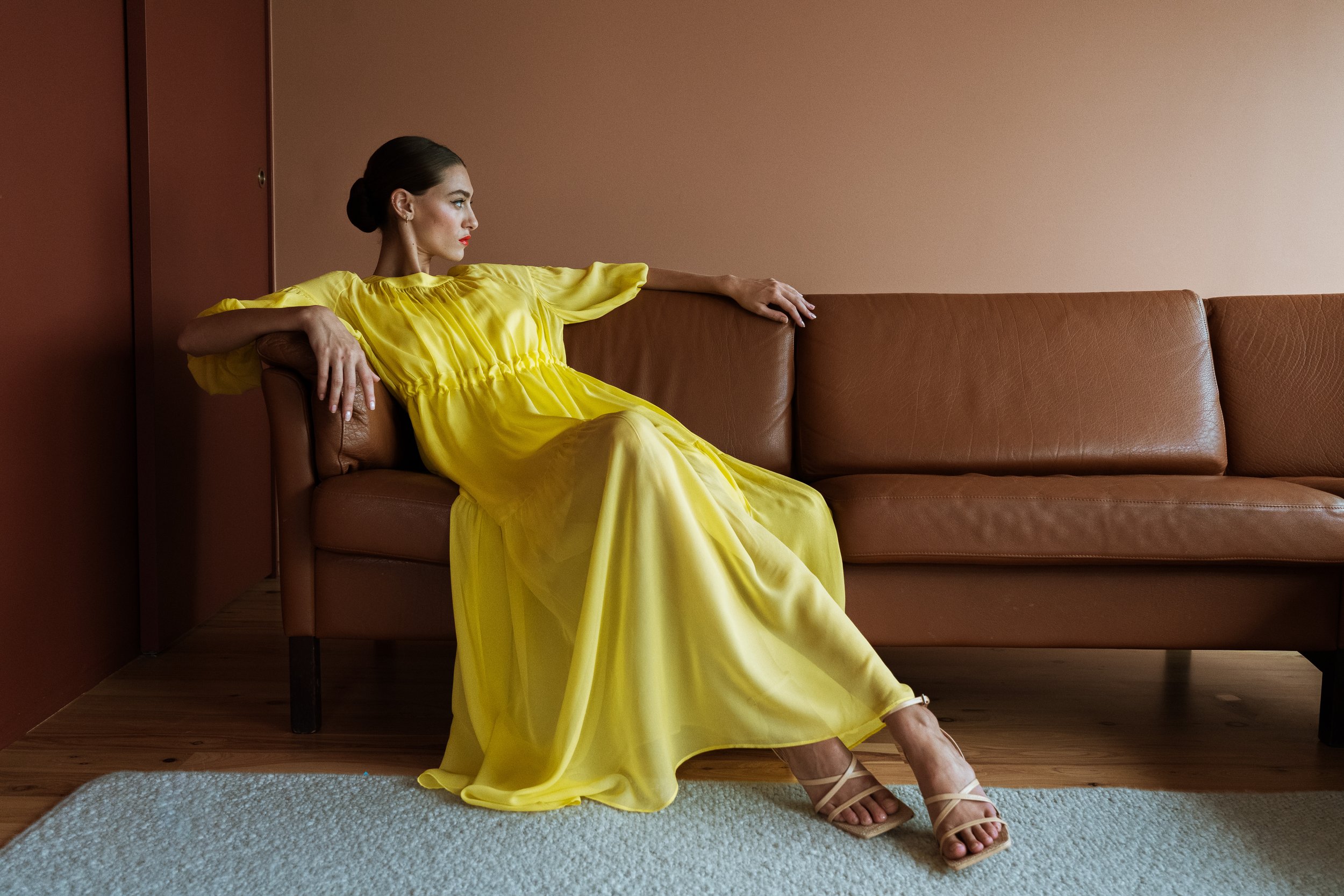 A woman in a flowing yellow dress sitting on a leather couch against a brown wall with hardwood floor and a small beige rug.