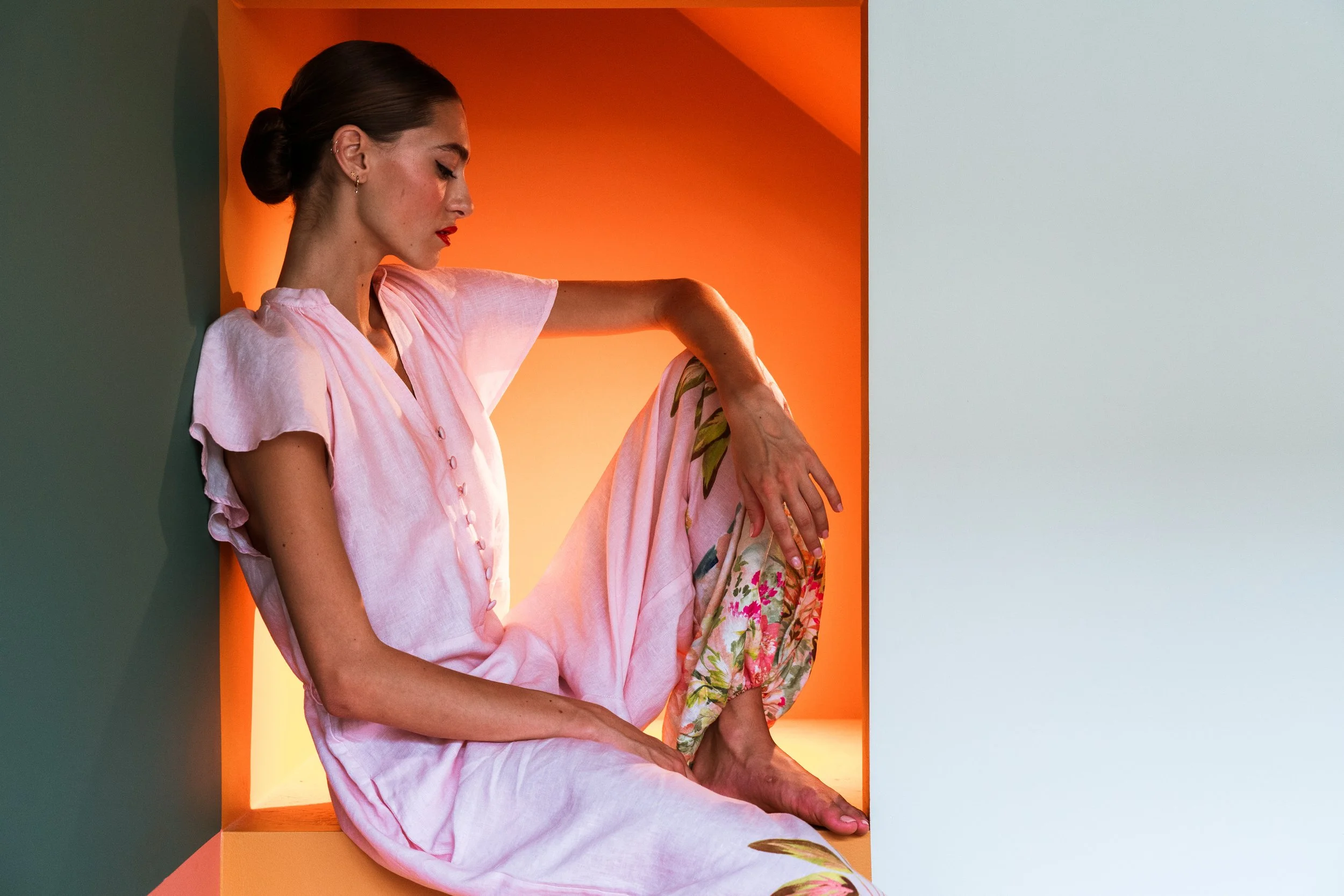 Woman with dark hair in a bun wearing a pink dress sitting inside a rectangular cutout with warm orange lighting, with a plain light-colored wall adjacent.